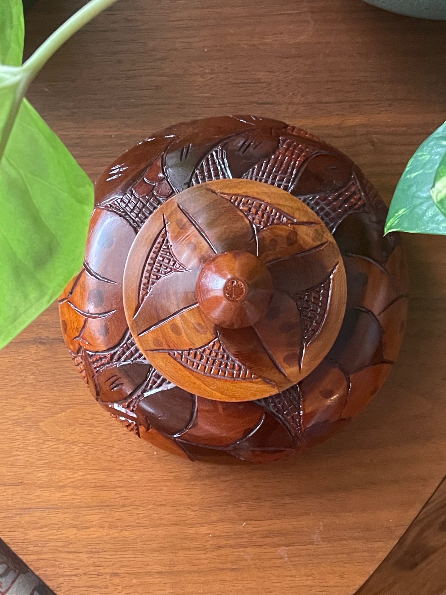 A round carved wooden box with a matching lid, featuring intricate floral and geometric engravings, placed on a wooden tabletop with green leaves visible at the edges.