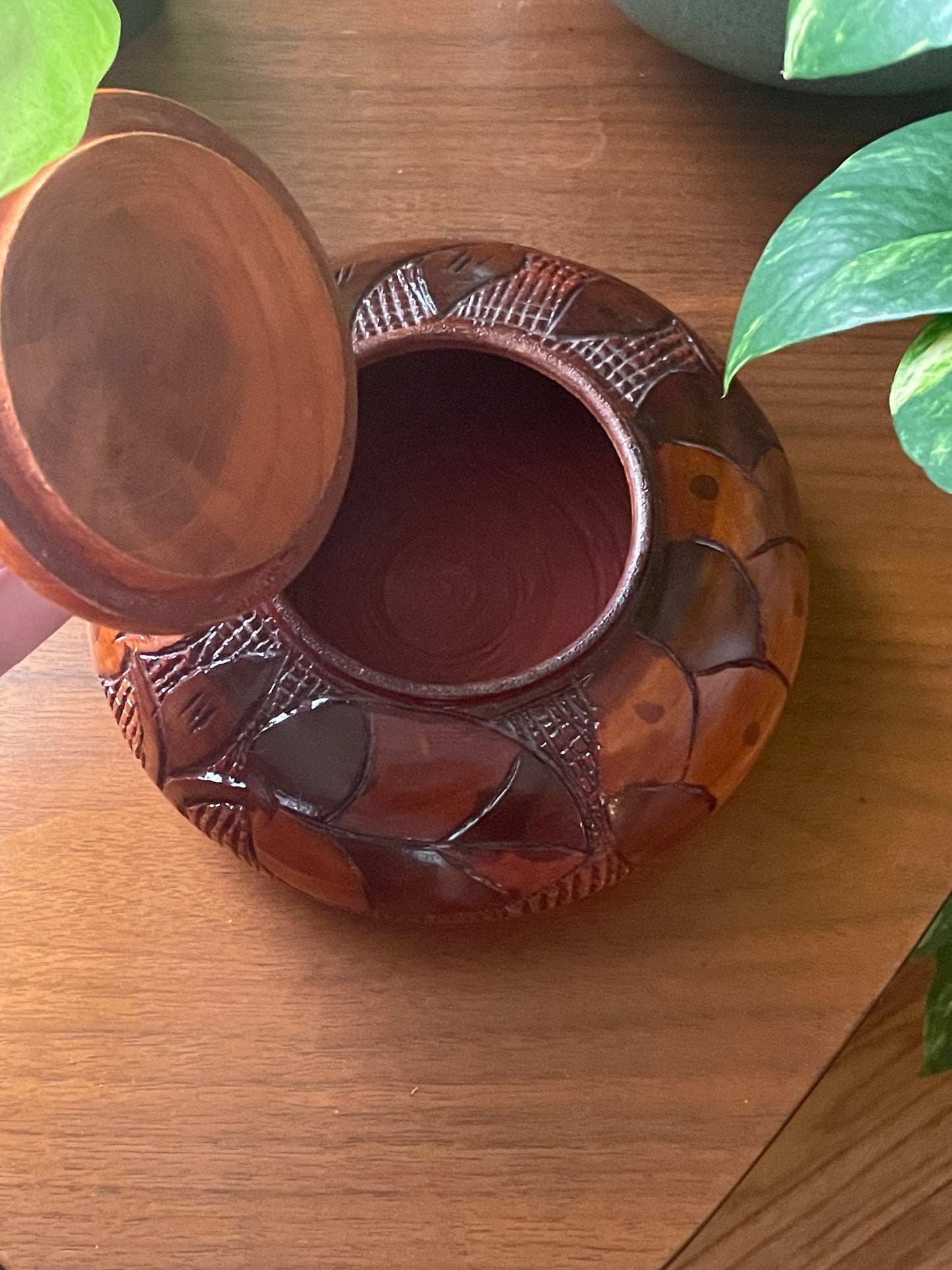 A carved wooden decorative box with a matching lid, shown on a wooden tabletop with green leaves in the background.