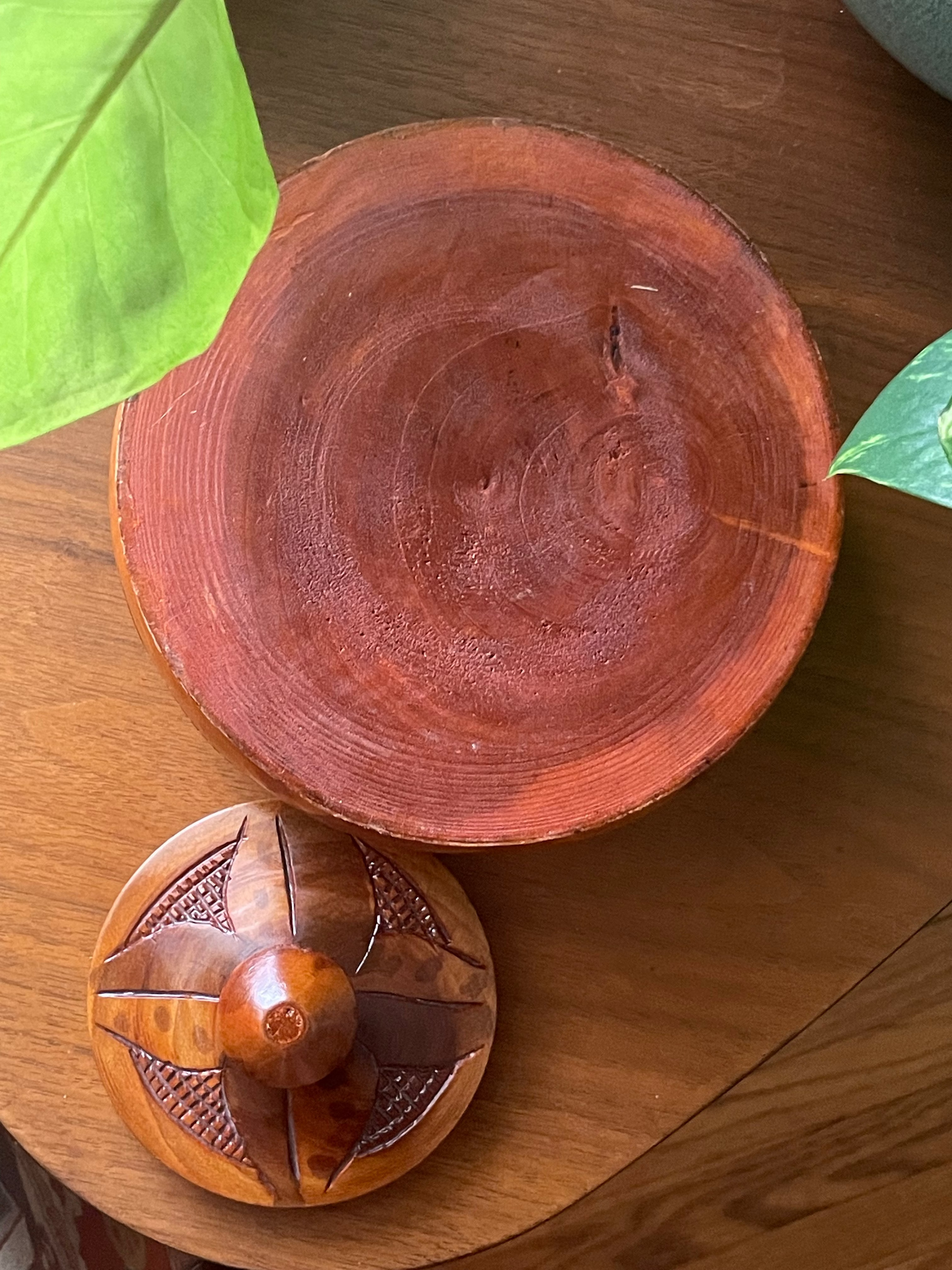 A handcrafted wooden box with a matching carved wooden lid, displayed on a wooden table with green leaves in the background.