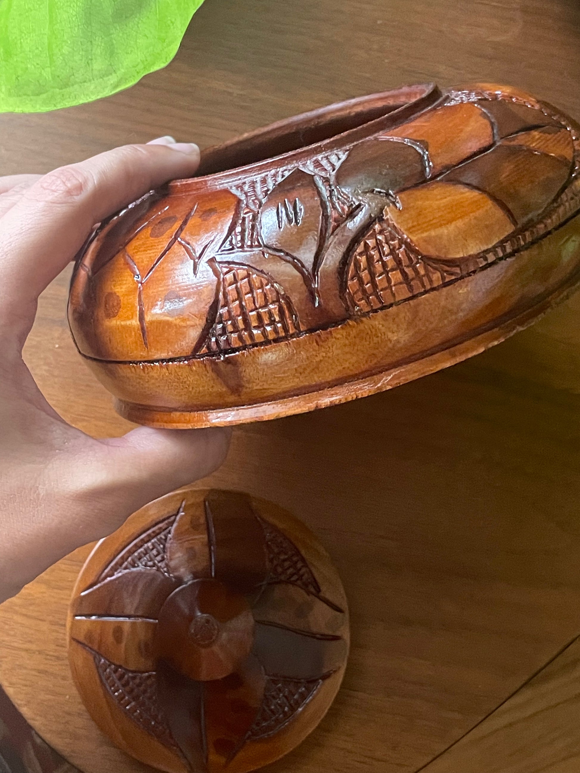 A hand holds a carved wooden box with a matching lid placed below it on a wooden surface. The box has an ornate floral and geometric pattern carved into the wood, showing warm brown and amber tones.
