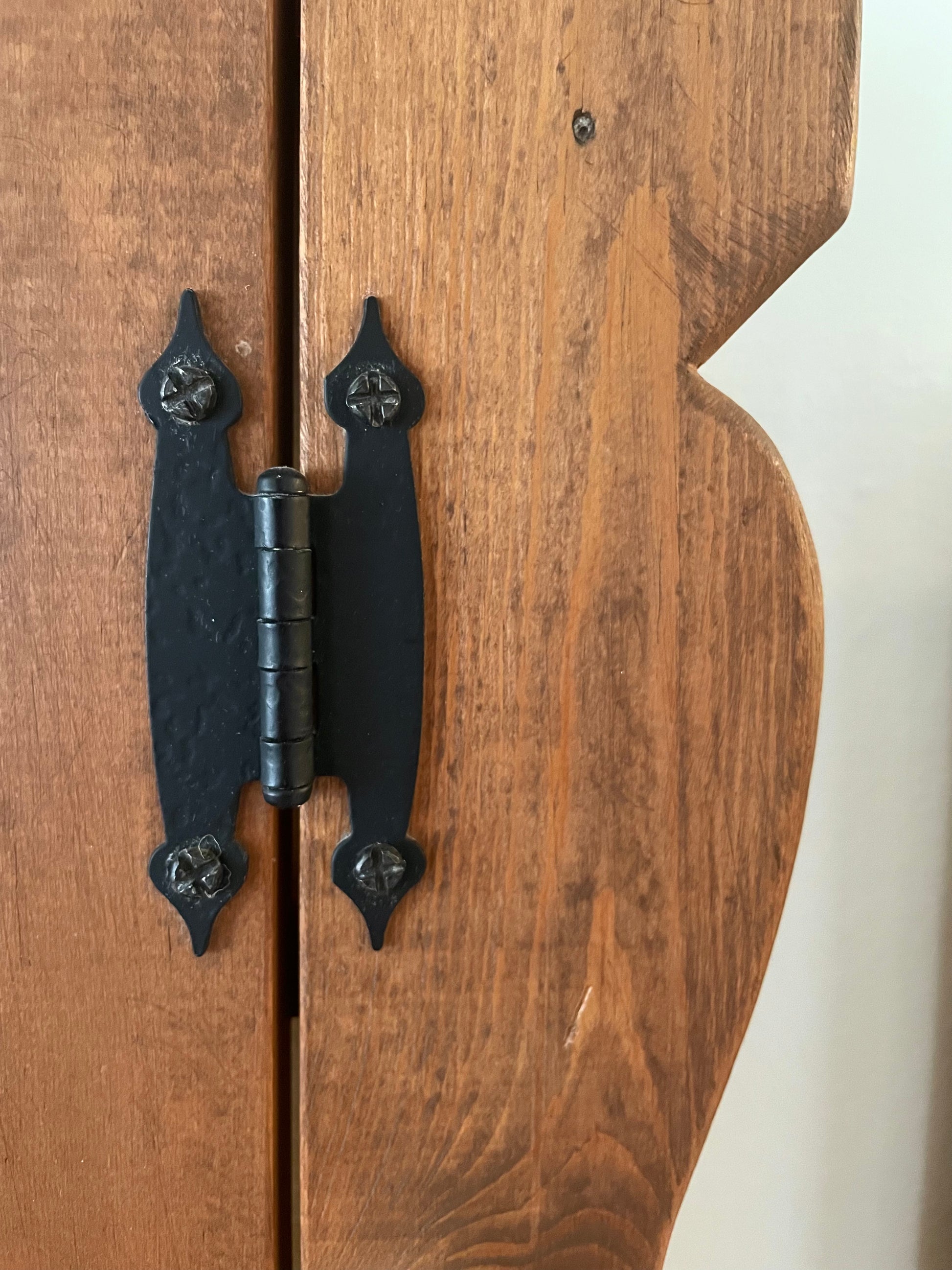 A close-up of a rustic black metal cabinet hinge attached to a warm brown wooden door with a scalloped edge.