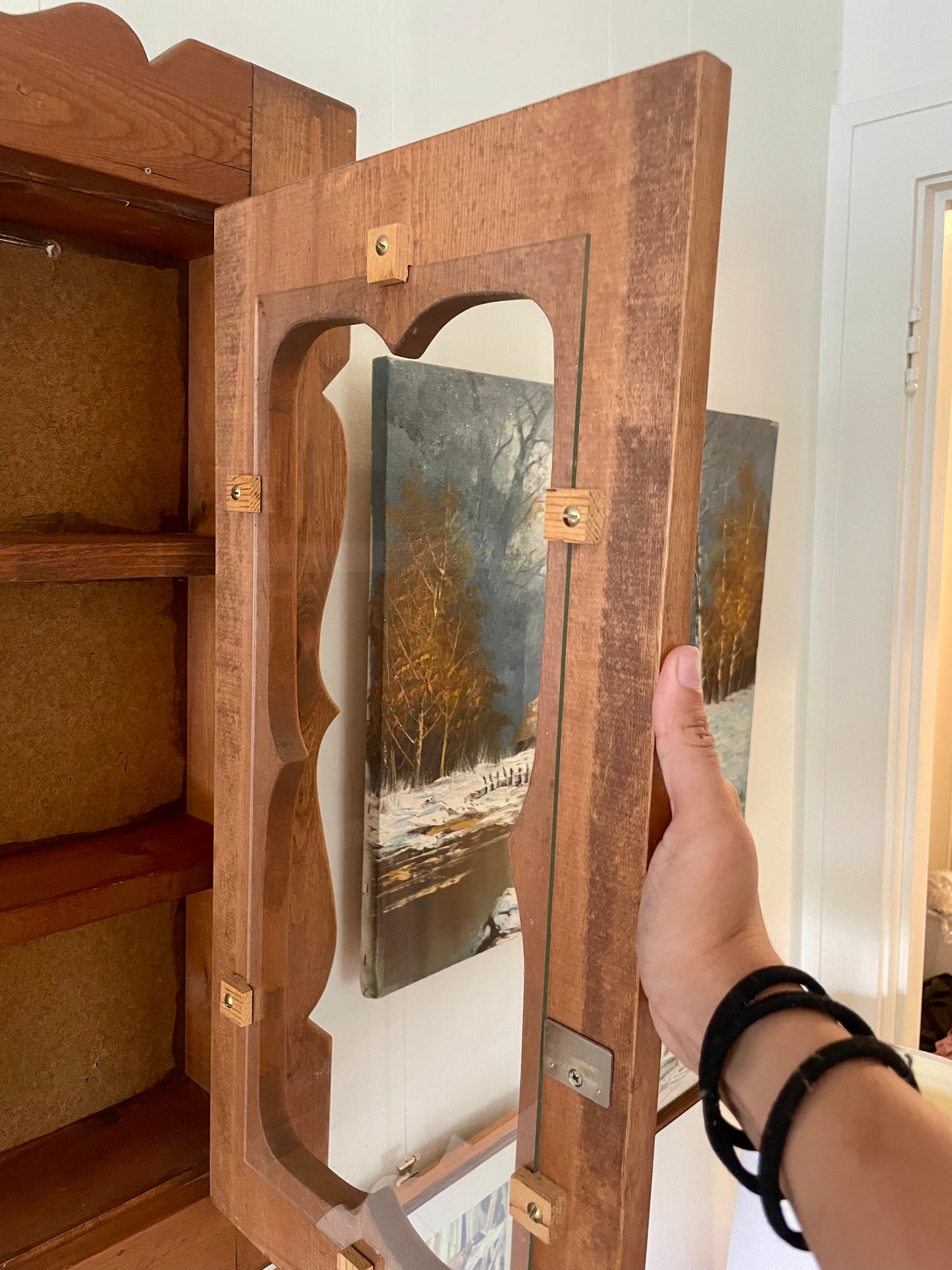 A hand holding a wooden cabinet door with scalloped cutout open, showing a winter forest landscape painting behind it.