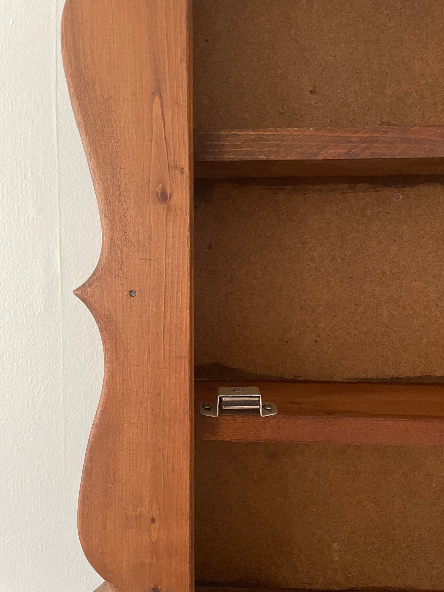 Close-up of a wooden cabinet door with a scalloped edge and two interior shelves, showing the warm medium-brown wood grain, a silver magnetic latch, and the brown particle board backing.