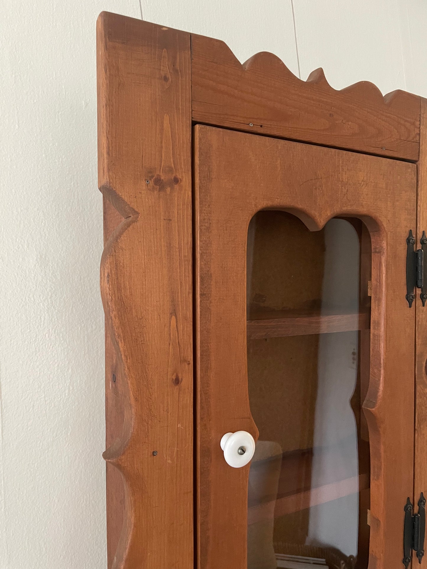 A close-up of a wooden cabinet with a warm medium-brown finish, featuring a glass-front door with a white round knob and decorative wavy trim along the top and sides.