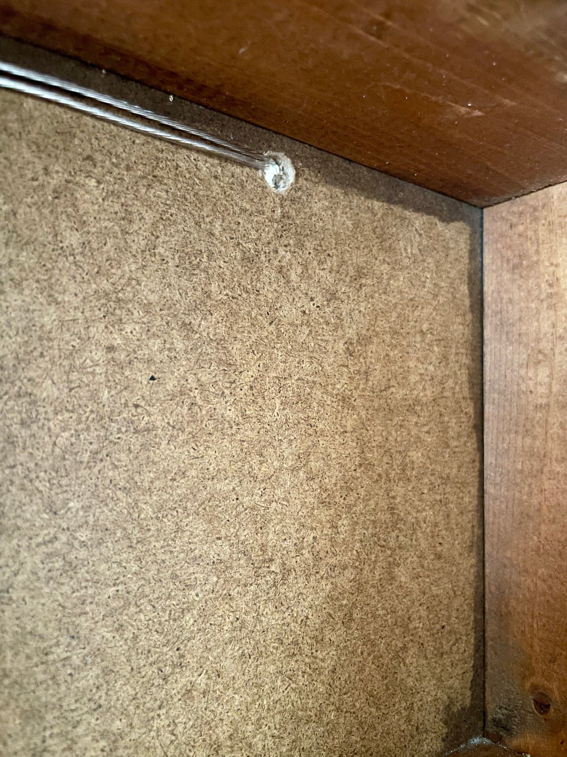 Close-up of the interior corner of a wooden wall cabinet, showing a brown textured particle board back panel, a wooden side panel, and a metal screw hole near the top.