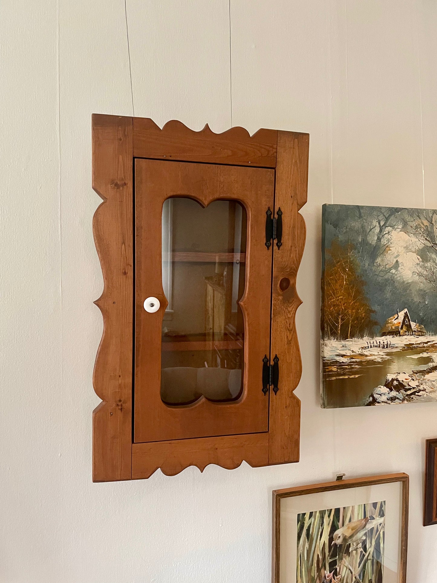 A rustic wooden wall cabinet with a scalloped frame and a glass door, mounted on a white paneled wall next to a landscape painting.