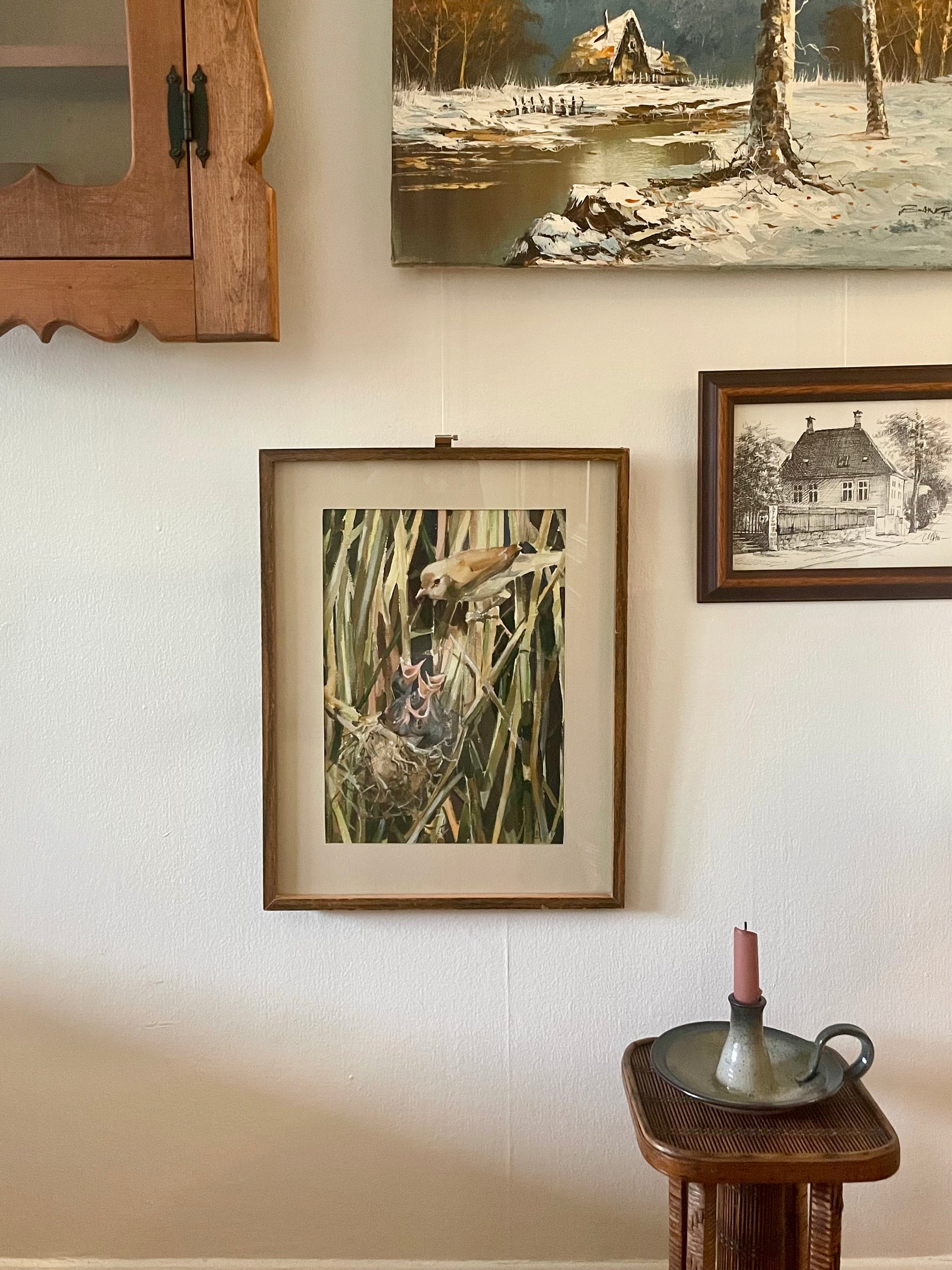 A framed bird-and-nest painting hangs on a beige wall in a styled interior setting, alongside other framed artwork, a wooden cabinet edge, and a small table with a candle holder.