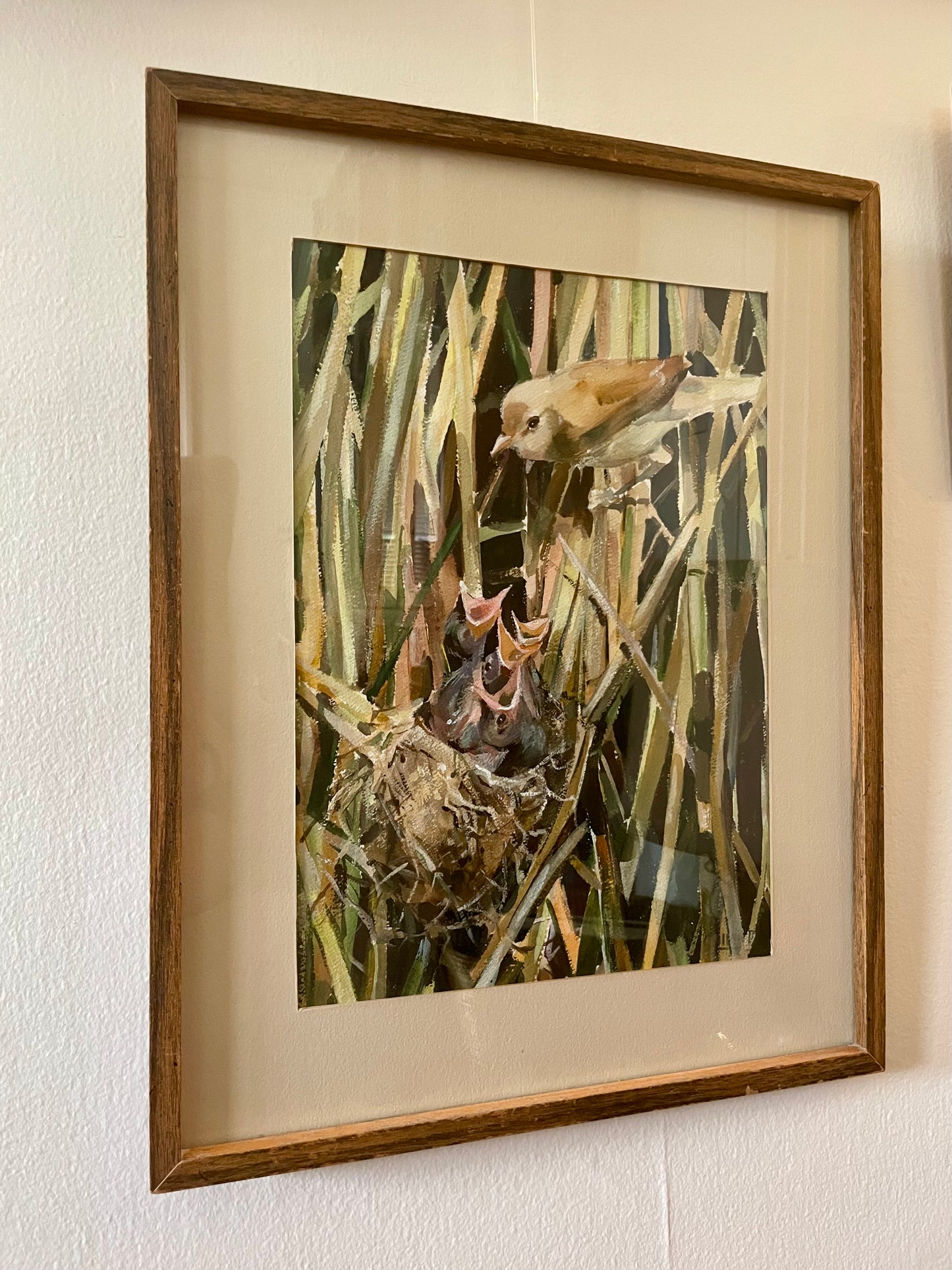 A framed painting depicting a small brown bird perched above a bird's nest with open beaks, mounted on a cream wall in a wooden frame with a beige mat.