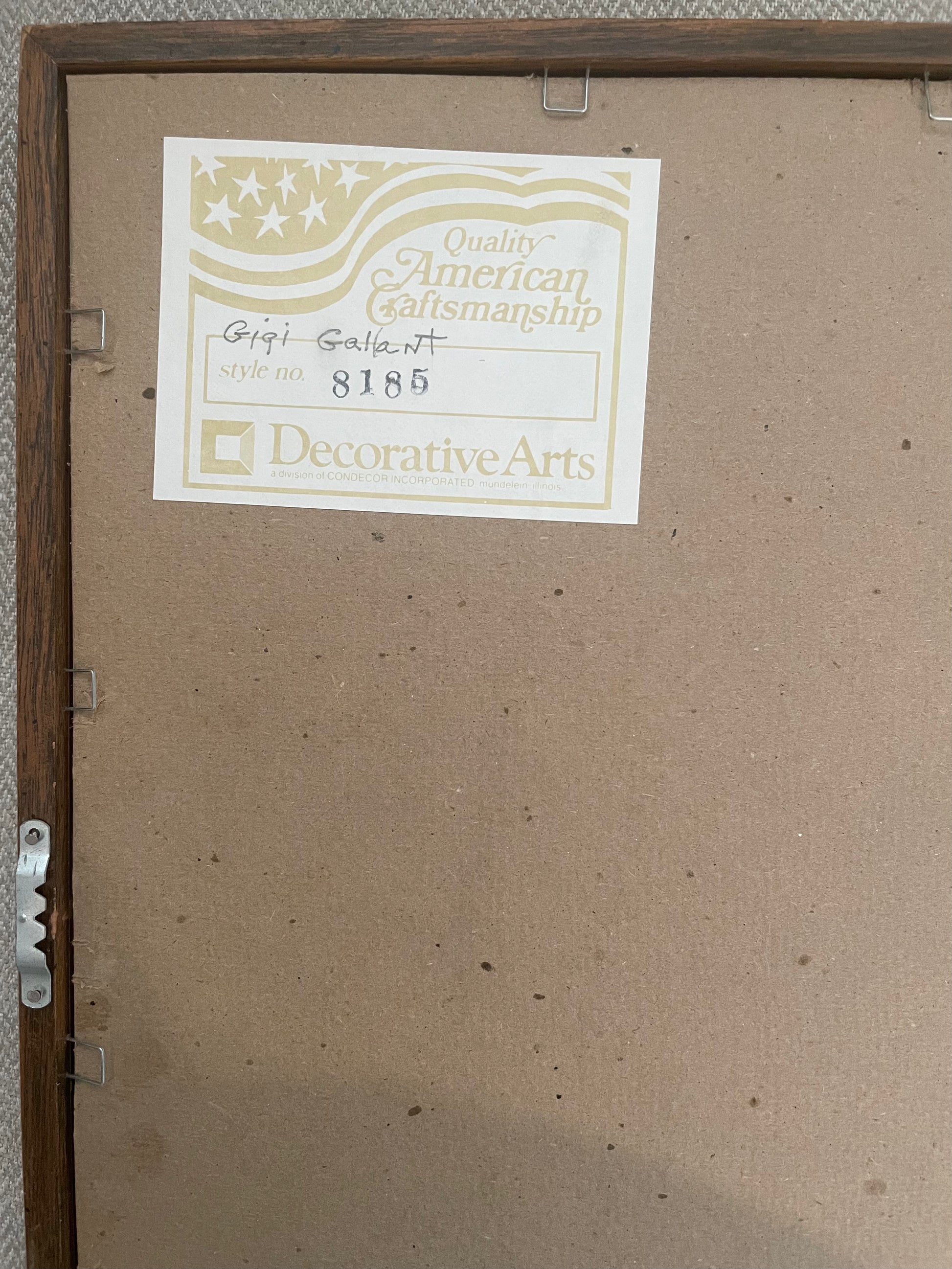 The back of a framed art piece showing a brown cardboard backing, a wooden frame, and a white paper label with 'Quality American Craftsmanship' and 'Decorative Arts' printed on it, signed with the artist’s name Gigi Gallant.