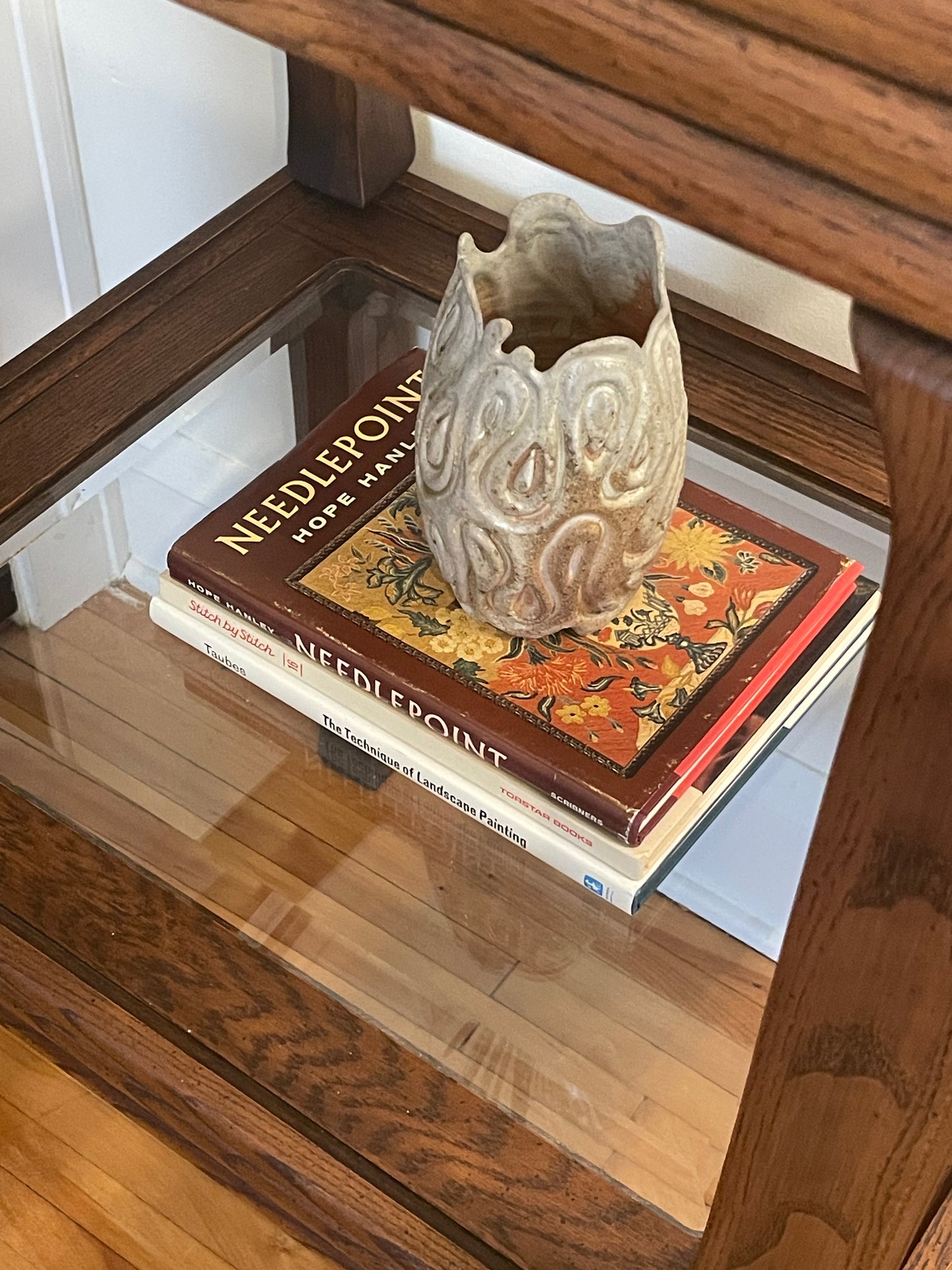 A small sculptural ceramic vase in a toasty beige color with a carved organic pattern, displayed on top of a stack of craft books inside a wooden table shelf.