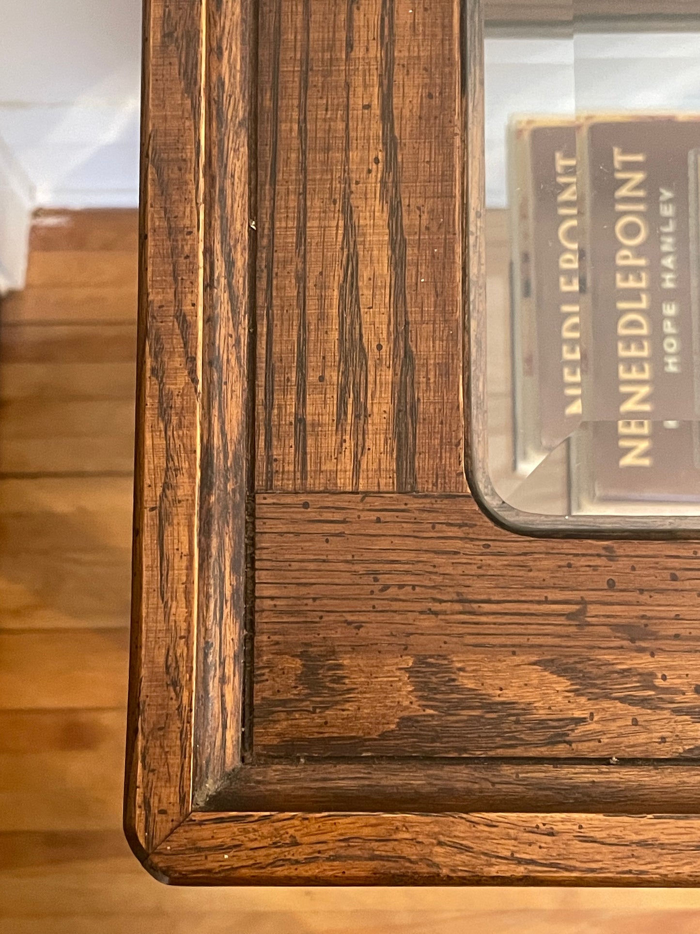 Close-up from the top of a carved wooden table corner with a glass top and rich medium-brown wood grain, showing a book titled Needlepoint by Hope Stanley beneath the glass.