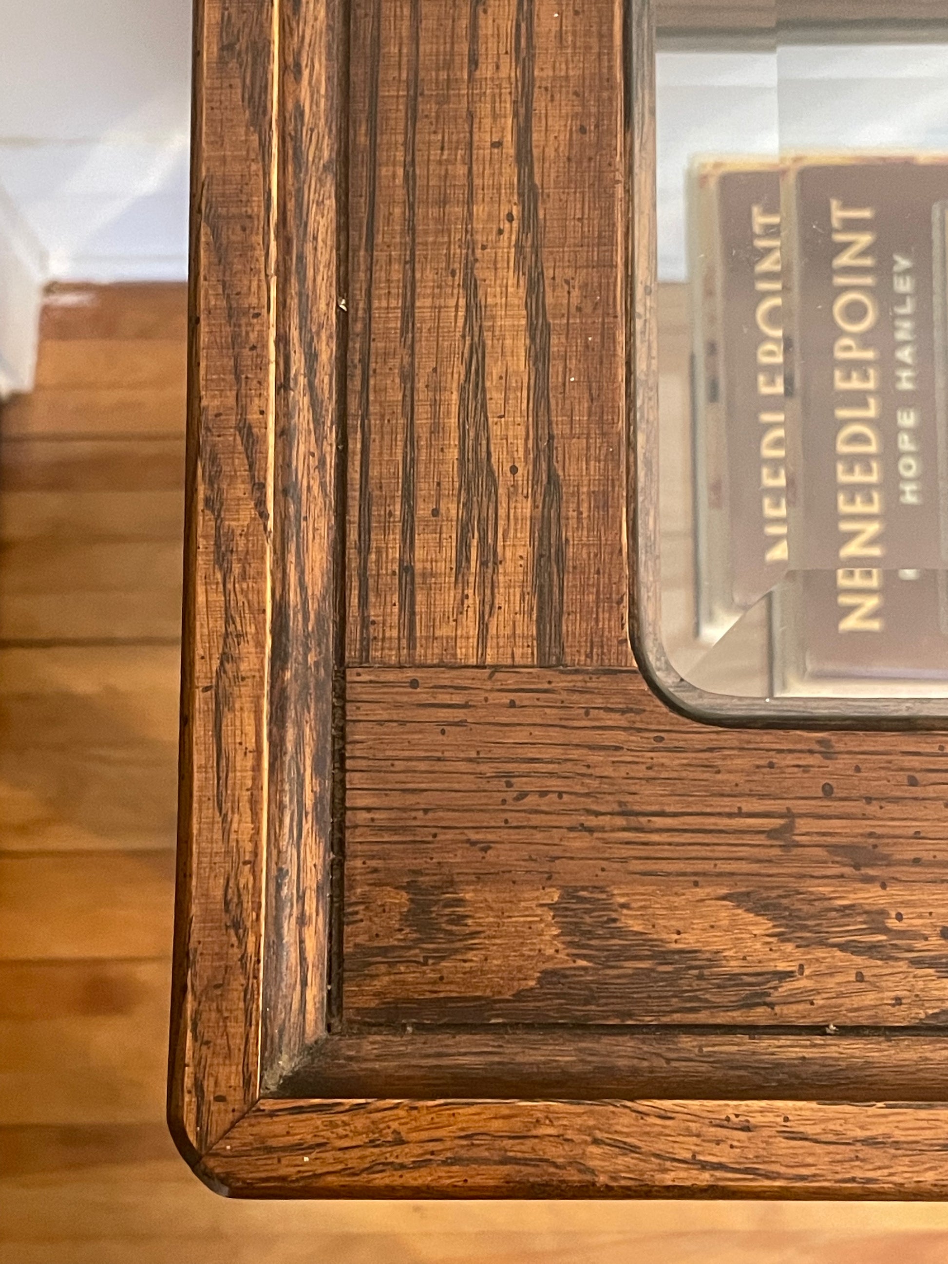 Close-up from the top of a carved wooden table corner with a glass top and rich medium-brown wood grain, showing a book titled Needlepoint by Hope Stanley beneath the glass.