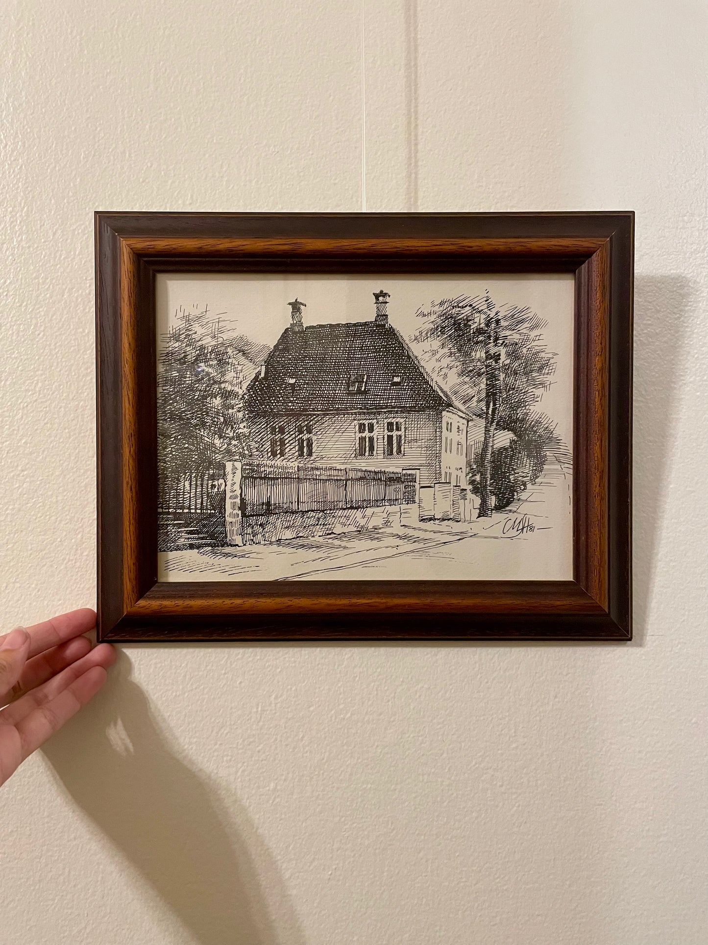A pen and ink drawing of a house on a tree-lined street, framed in a duo-toned wood frame and mounted on a textured off-white wall. A hand is visible on the left holding the frame.