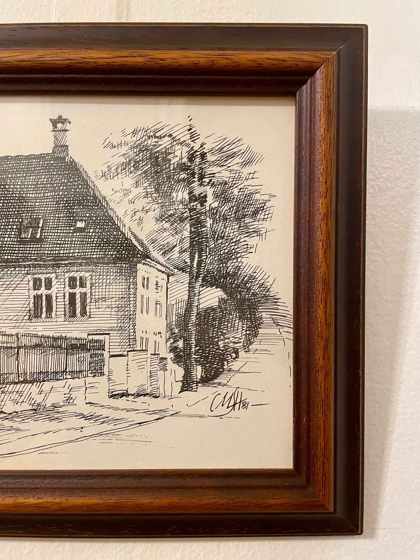 Closeup of a pen and ink drawing of a house on a tree-lined street, framed in a duo-toned wood frame and mounted on a textured off-white wall. 