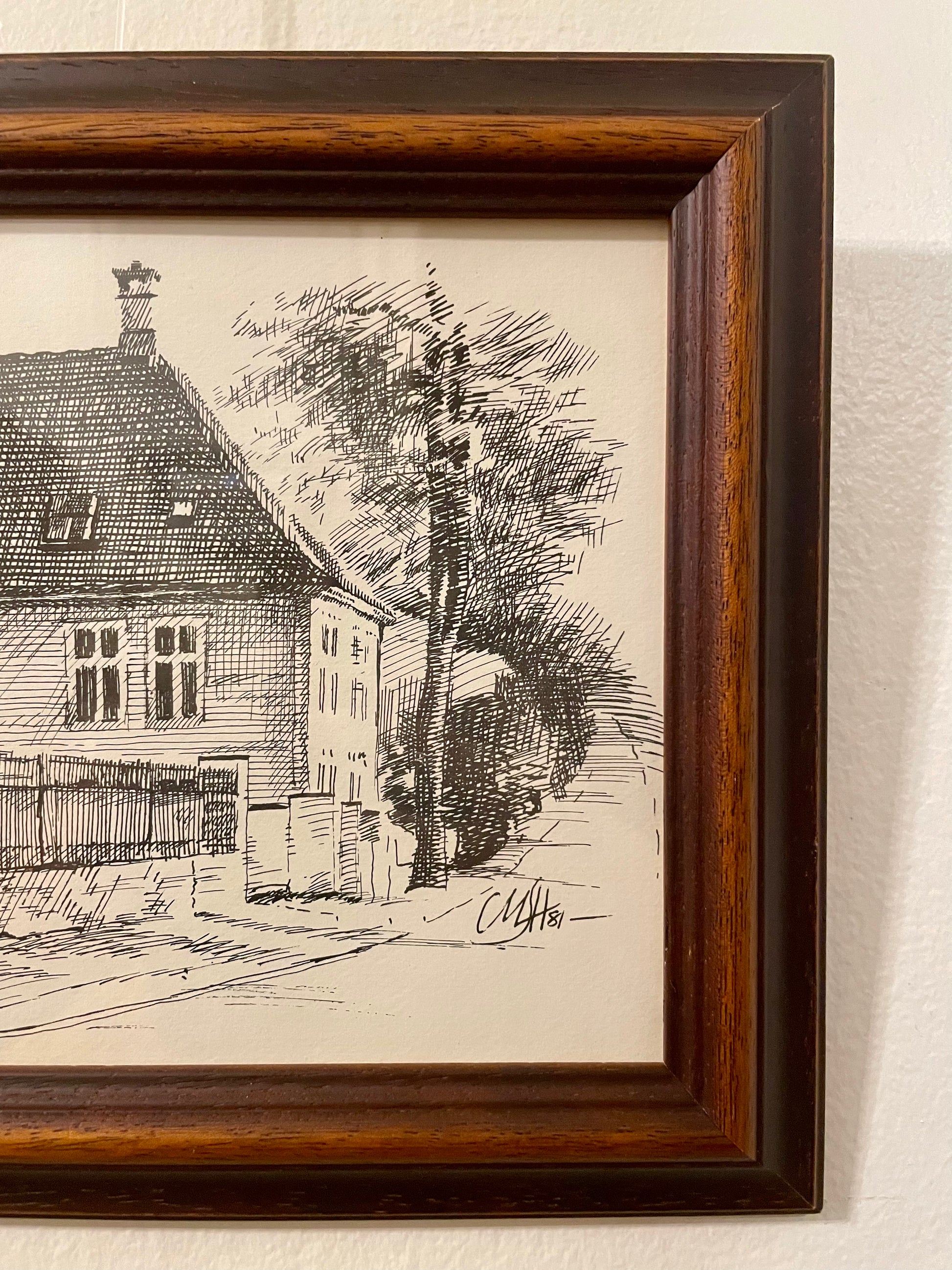 Closeup of a pen and ink drawing of a house on a tree-lined street, framed in a duo-toned wood frame and mounted on a textured off-white wall. 