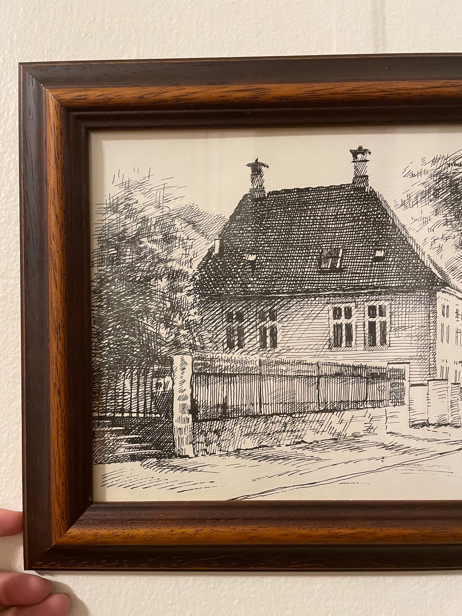 Closeup of a pen and ink drawing of a house on a tree-lined street, framed in a duo-toned wood frame and mounted on a textured off-white wall. A hand is visible holding the frame at the left.