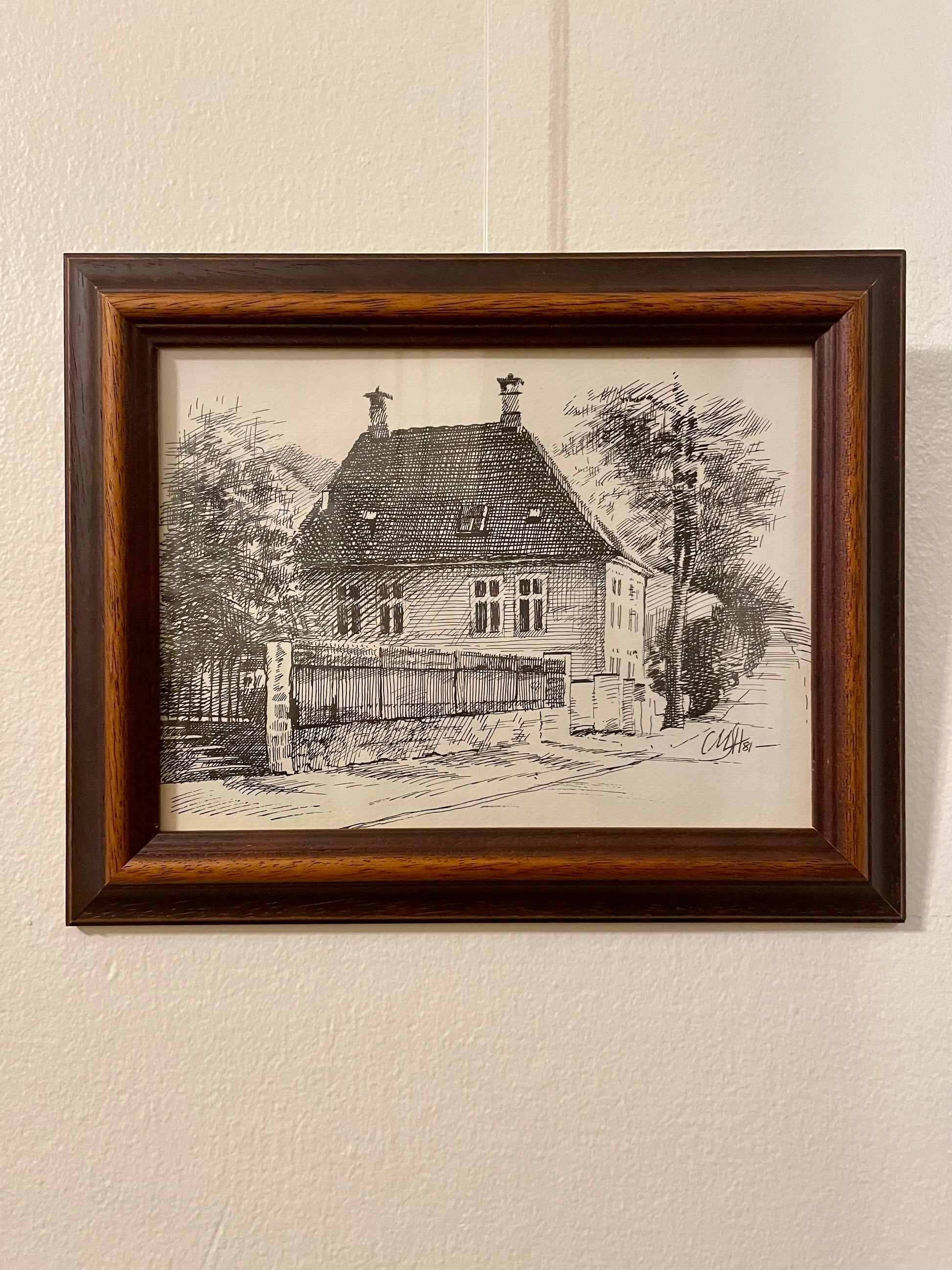 Pen and ink drawing of a house on a tree-lined street, framed in a duo-toned wood frame and mounted on a textured off-white wall. 