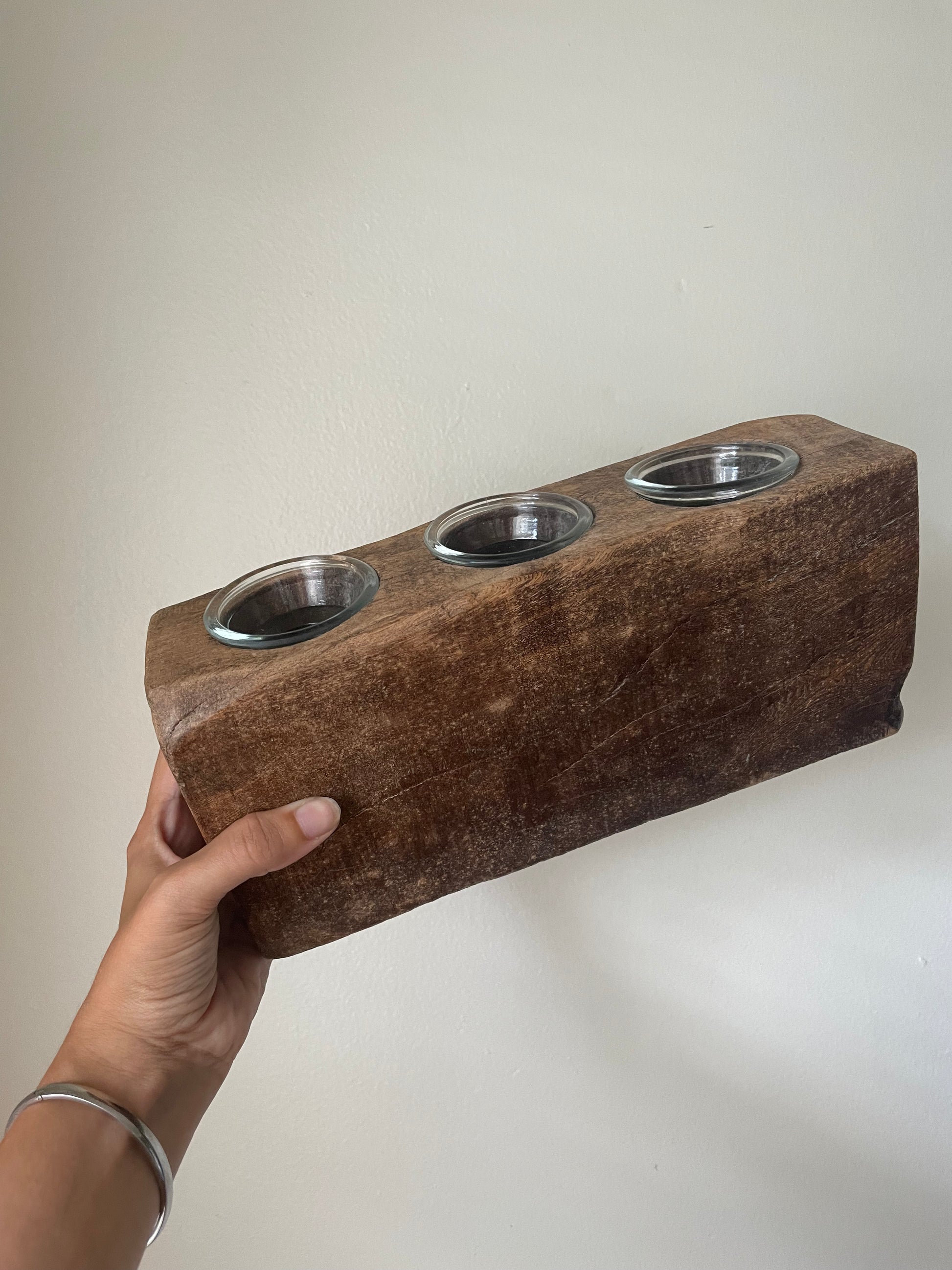A hand holding a rustic dark brown wooden block with three inset circular glass votives, set against a plain off-white wall.