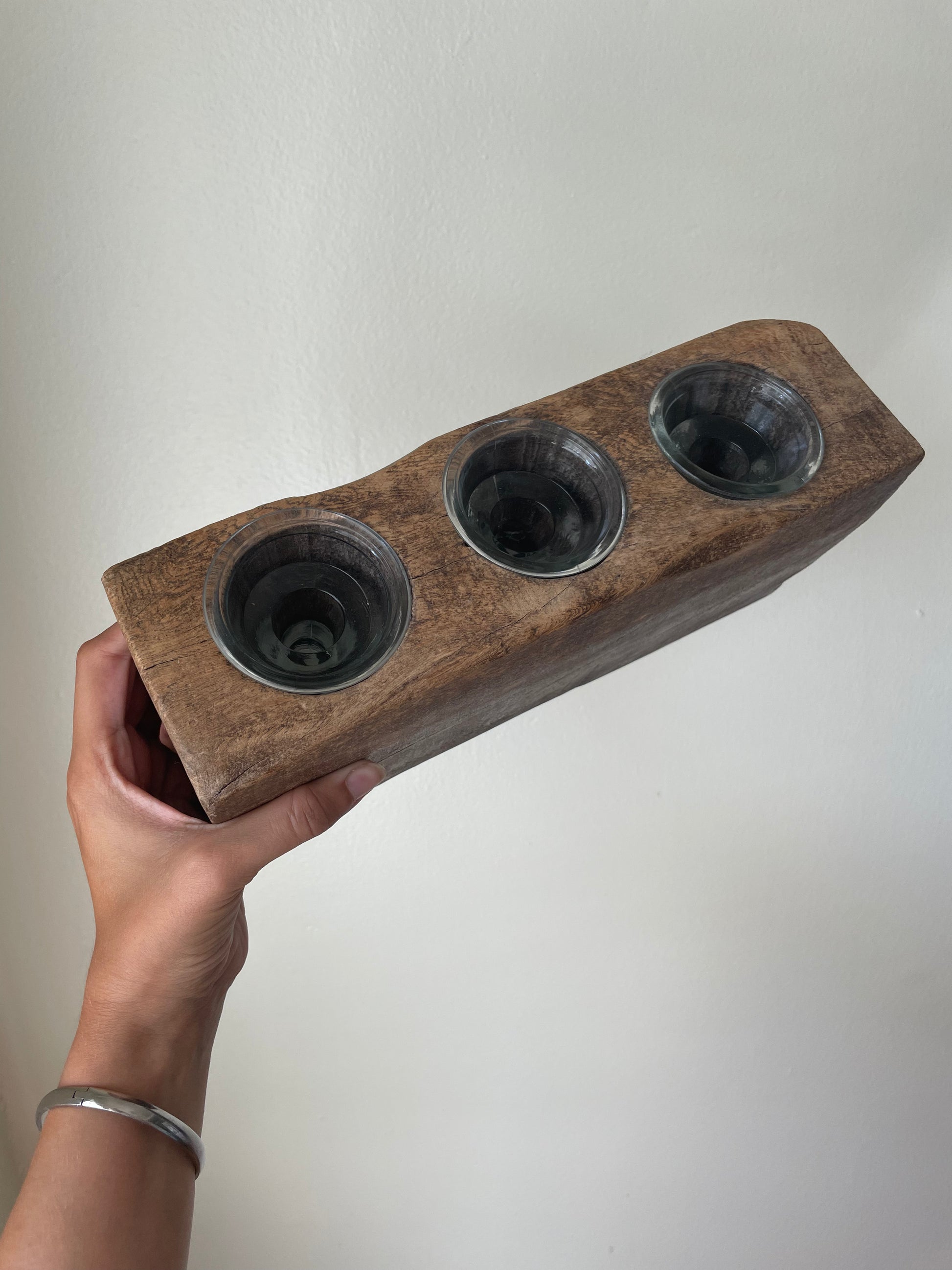 A hand holding a rustic brown wooden block with three inset circular glass votives, set against a plain off-white wall.