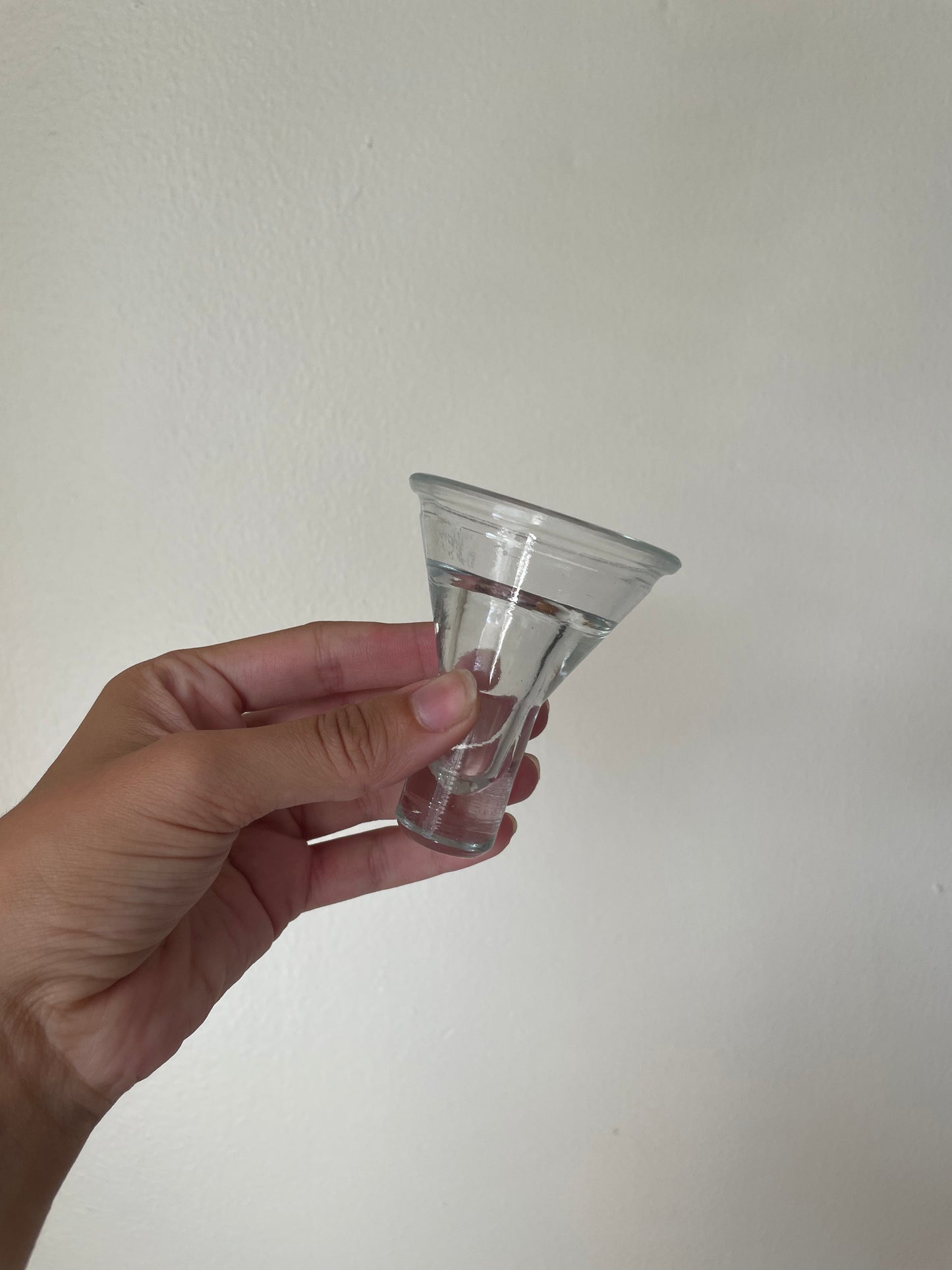 A hand holding a small clear glass cone-shaped votive against a plain off-white wall background.