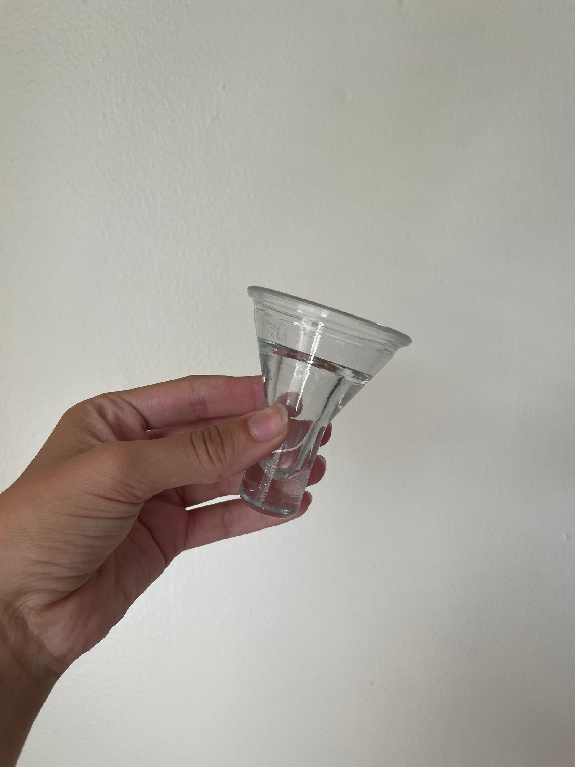 A hand holding a small clear glass cone-shaped votive against a plain off-white wall background.