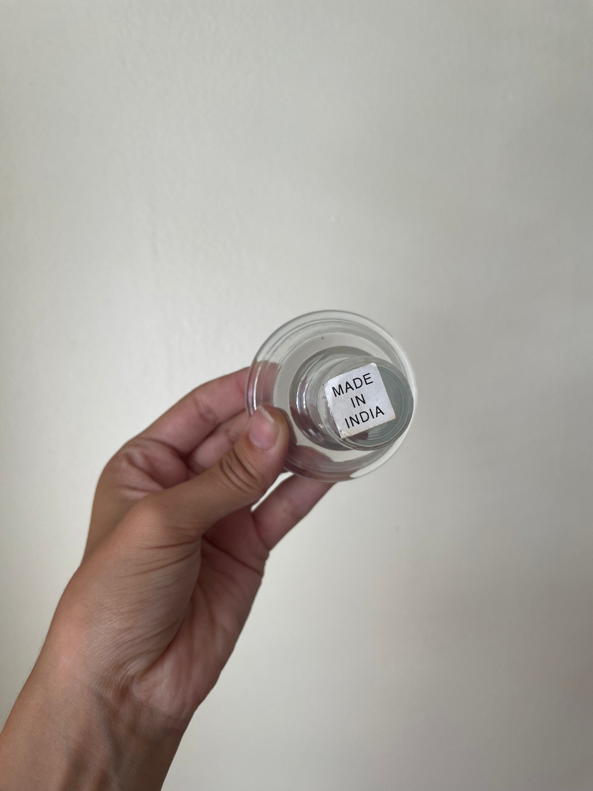 A hand holding a small clear round glass votive with a white label that reads 'MADE IN INDIA' against a plain off-white wall background.