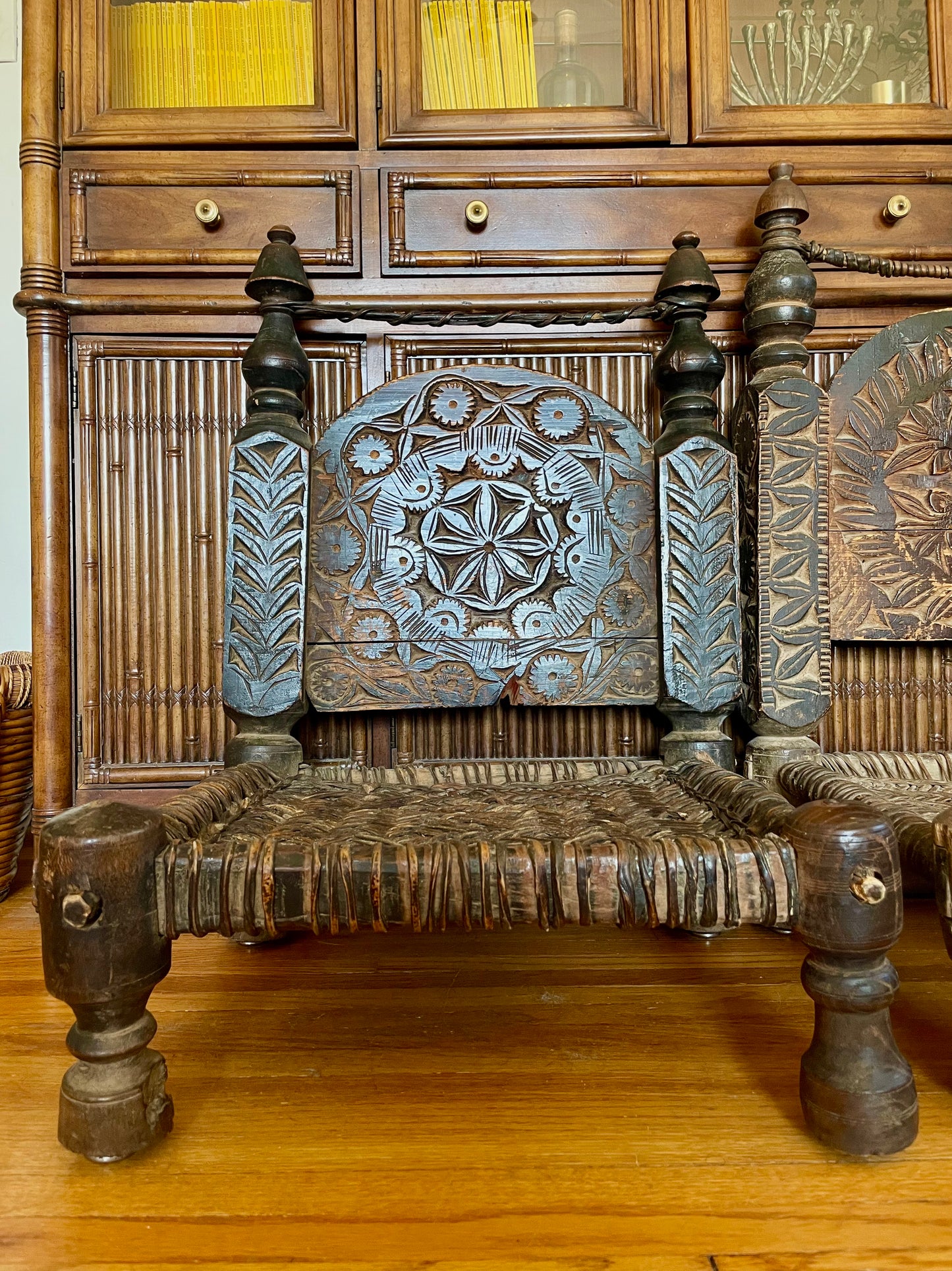 An ornate carved wooden chair with a woven seat and low legs, positioned in front of a large wooden cabinet.