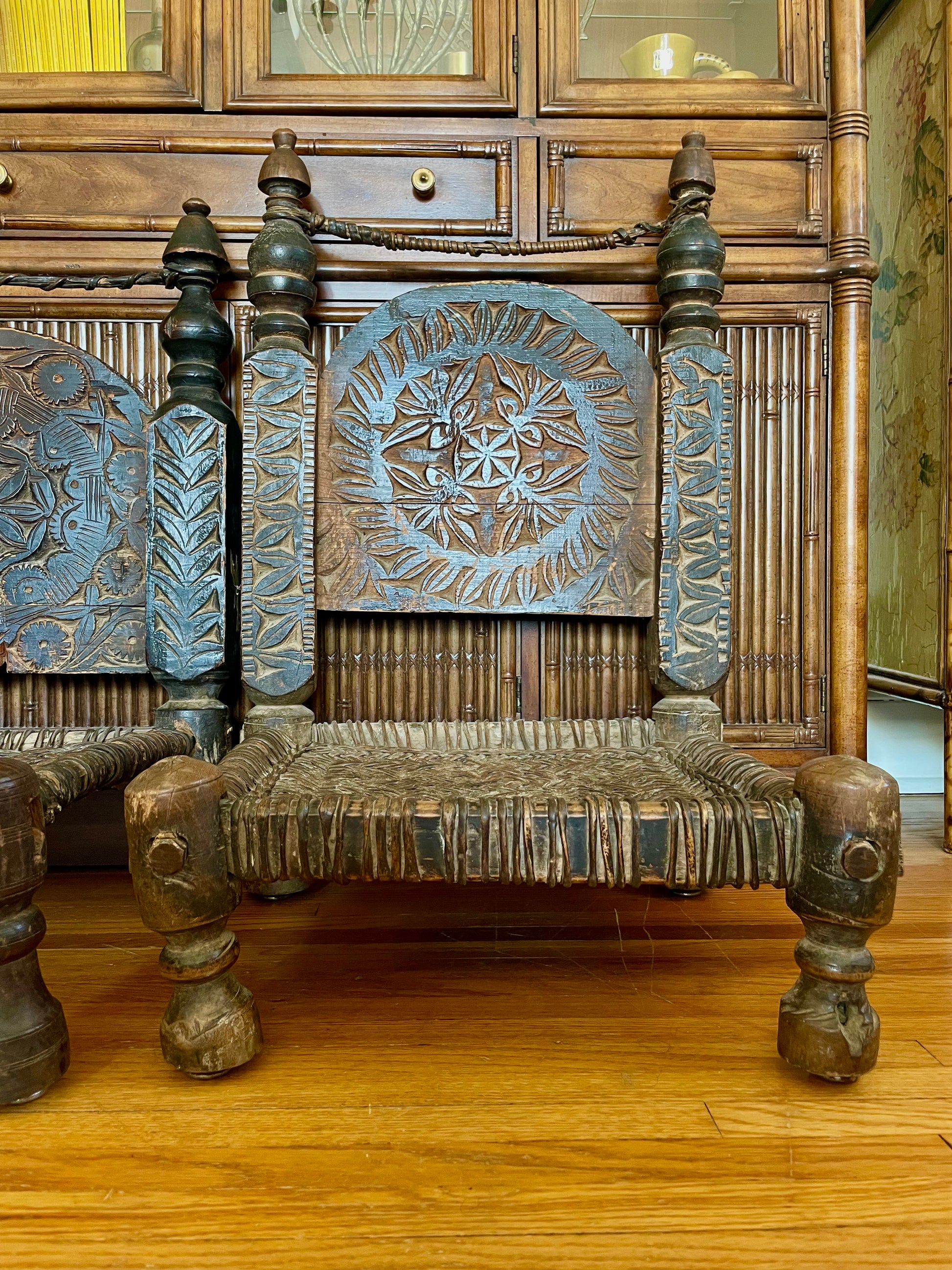 An ornate carved wooden chair with a woven seat and low legs, positioned in front of a large wooden cabinet.