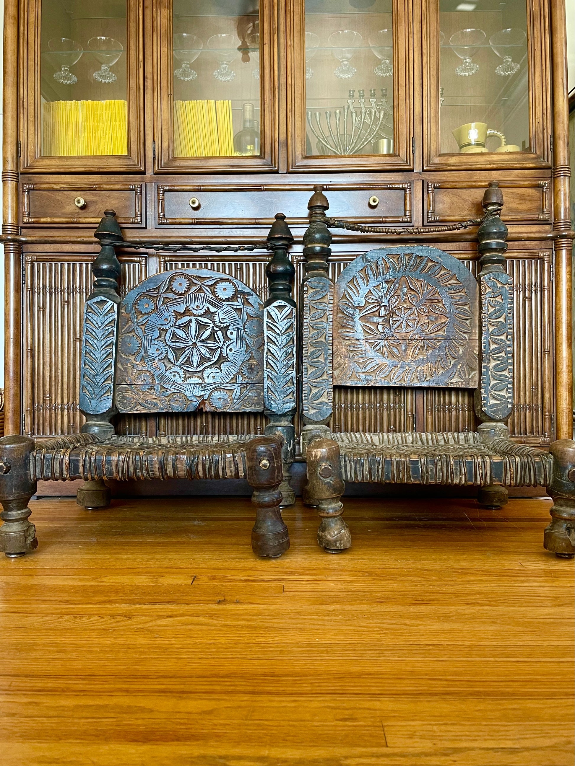 Two hand-carved wooden chairs with ornate floral and geometric designs on the backrests, positioned in front of a large wooden hutch on a polished hardwood floor.