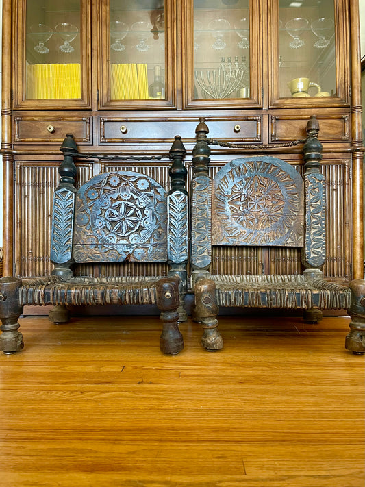Two hand-carved wooden chairs with ornate floral and geometric designs on the backrests, positioned in front of a large wooden hutch on a polished hardwood floor.
