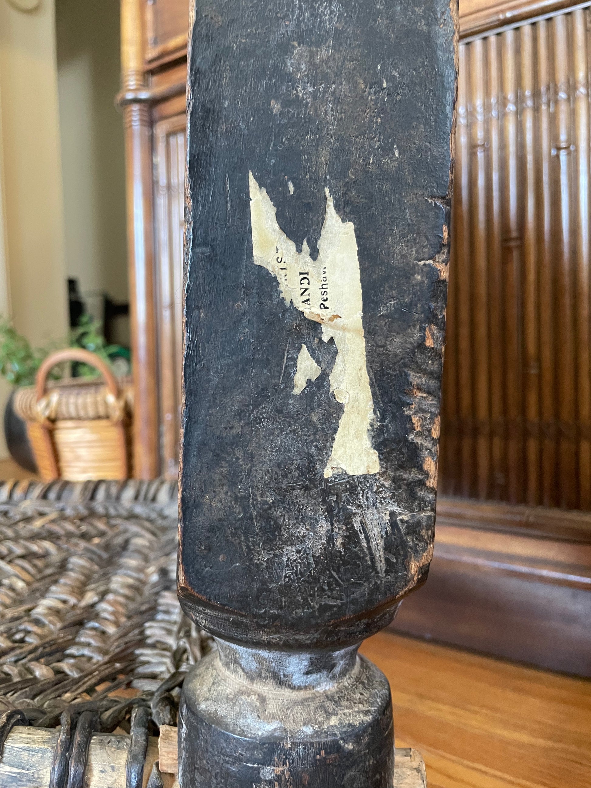 A close-up of the side of a weathered dark wooden chair showing an aged and peeling white adhesive label, set against a background of a wooden cabinet.
