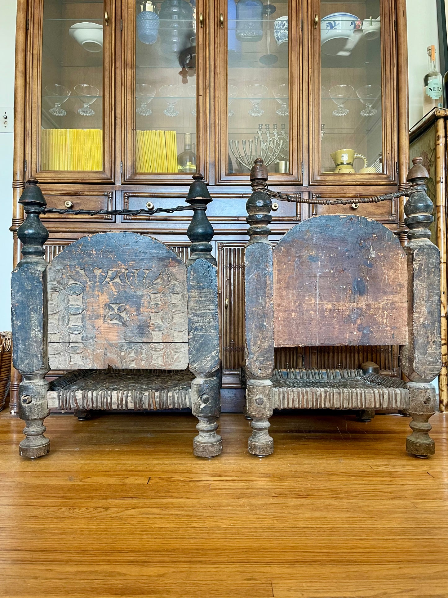 The backside of two wooden chairs with curved backs, distressed finish, woven seats, and short turned wood legs styled on a hardwood floor in front of a dark wood hutch.