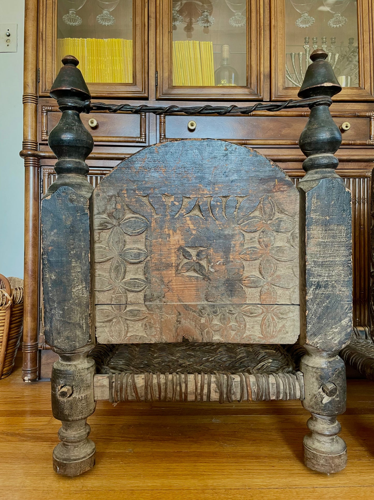 The backside of a wooden chair with a distressed brown finish and geometric carved design, featuring turned spindle legs, a woven seat, and a curved wooden backrest, positioned in front of a wooden hutch.