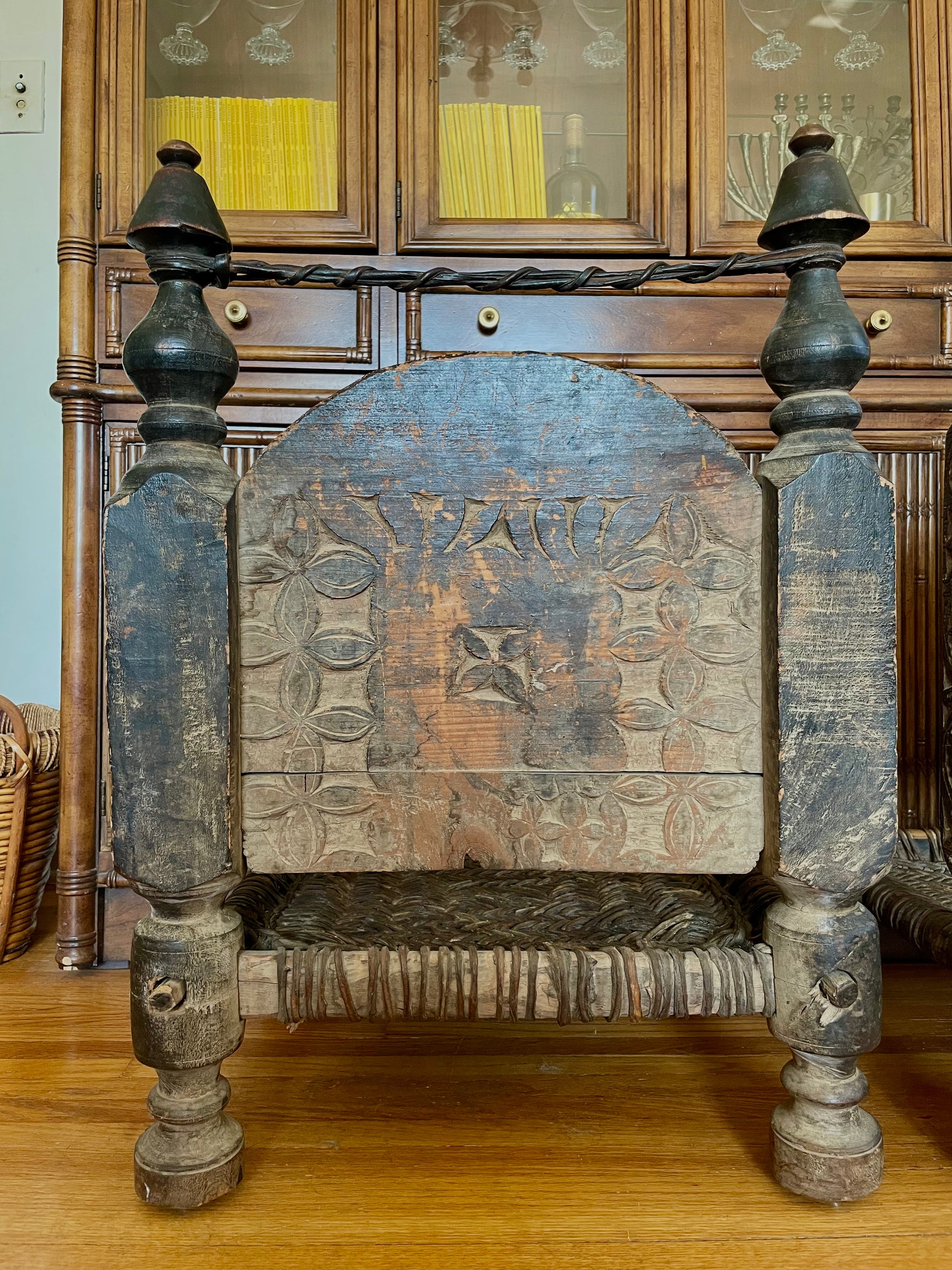 The backside of a wooden chair with a distressed brown finish and geometric carved design, featuring turned spindle legs, a woven seat, and a curved wooden backrest, positioned in front of a wooden hutch.