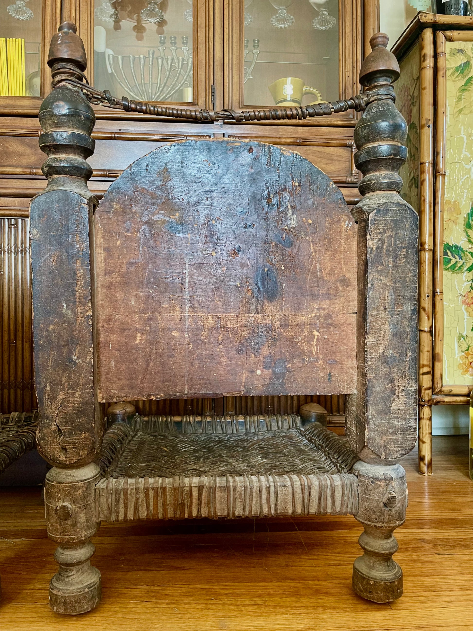 The backside of a wooden chair with a distressed brown finish, featuring turned spindle legs, a woven seat, and a curved wooden backrest, set against a background of a wooden cabinet and bamboo furniture.