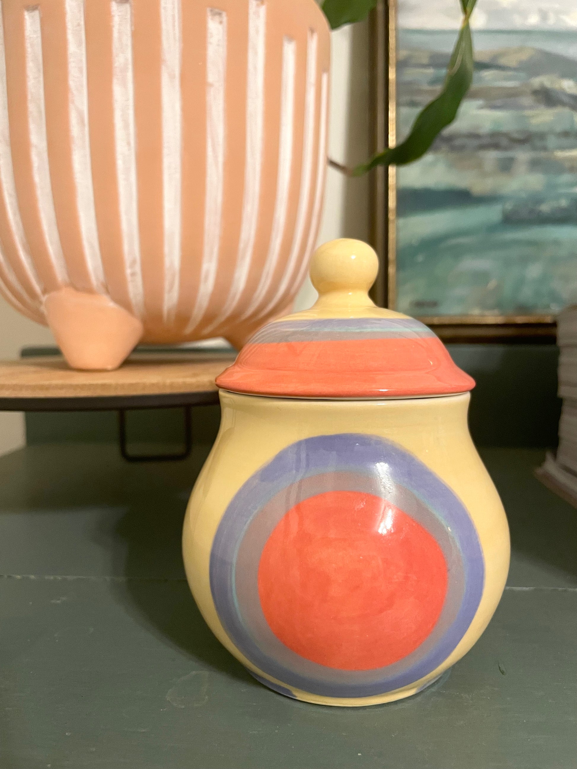 A ceramic lidded jar with a rounded body, featuring a hand-painted butter yellow base with concentric circles in coral, periwinkle, and lavender and a matching lid.