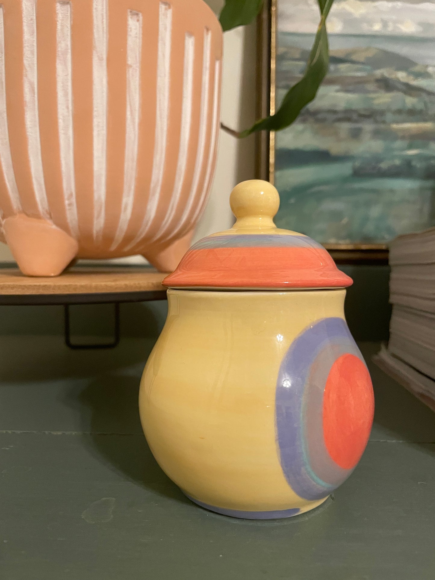 Side view of a ceramic lidded jar with a rounded body, featuring a hand-painted butter yellow base with concentric circles in coral, periwinkle, and lavender and a matching lid.