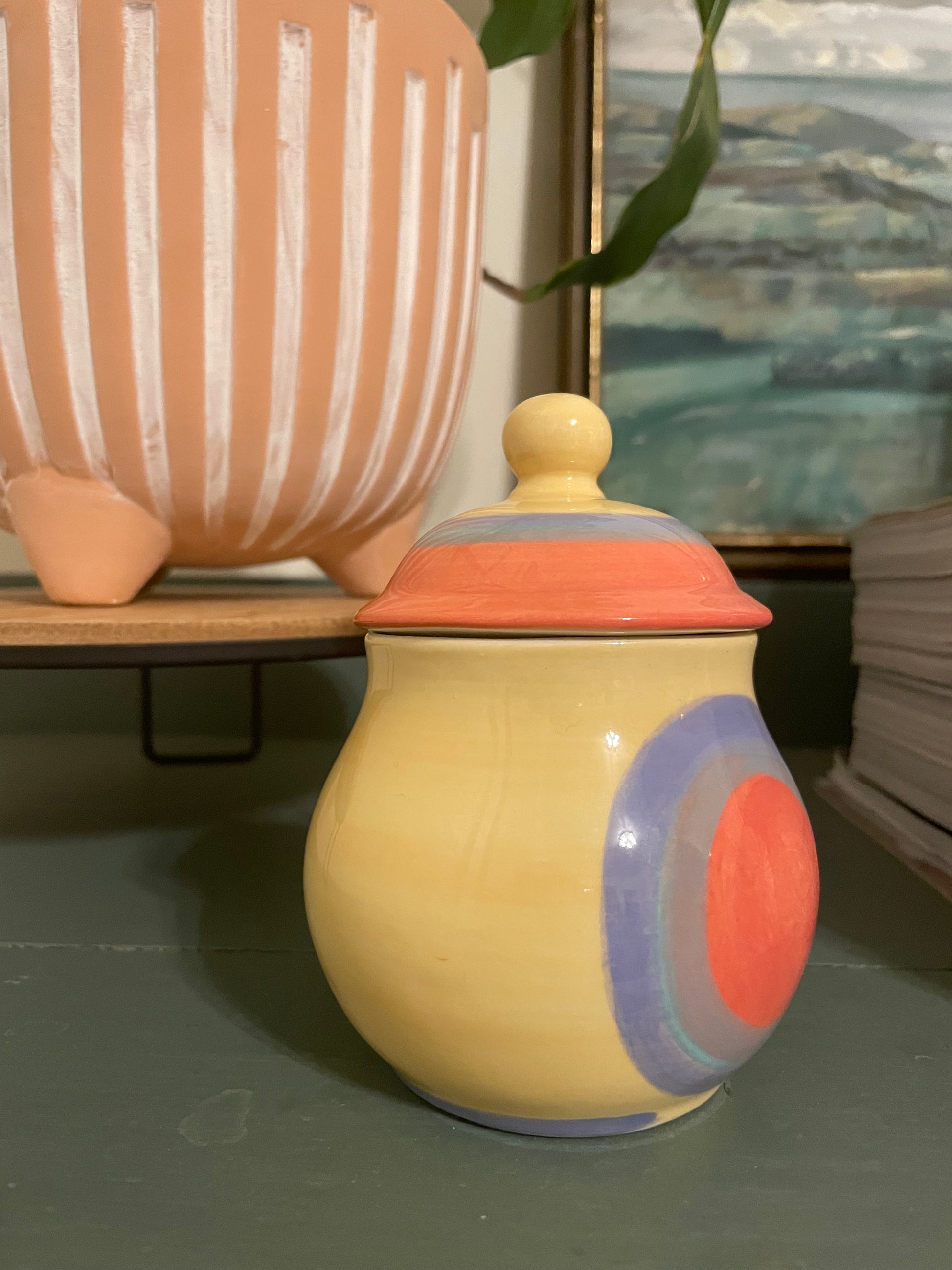 Side view of a ceramic lidded jar with a rounded body, featuring a hand-painted butter yellow base with concentric circles in coral, periwinkle, and lavender and a matching lid.