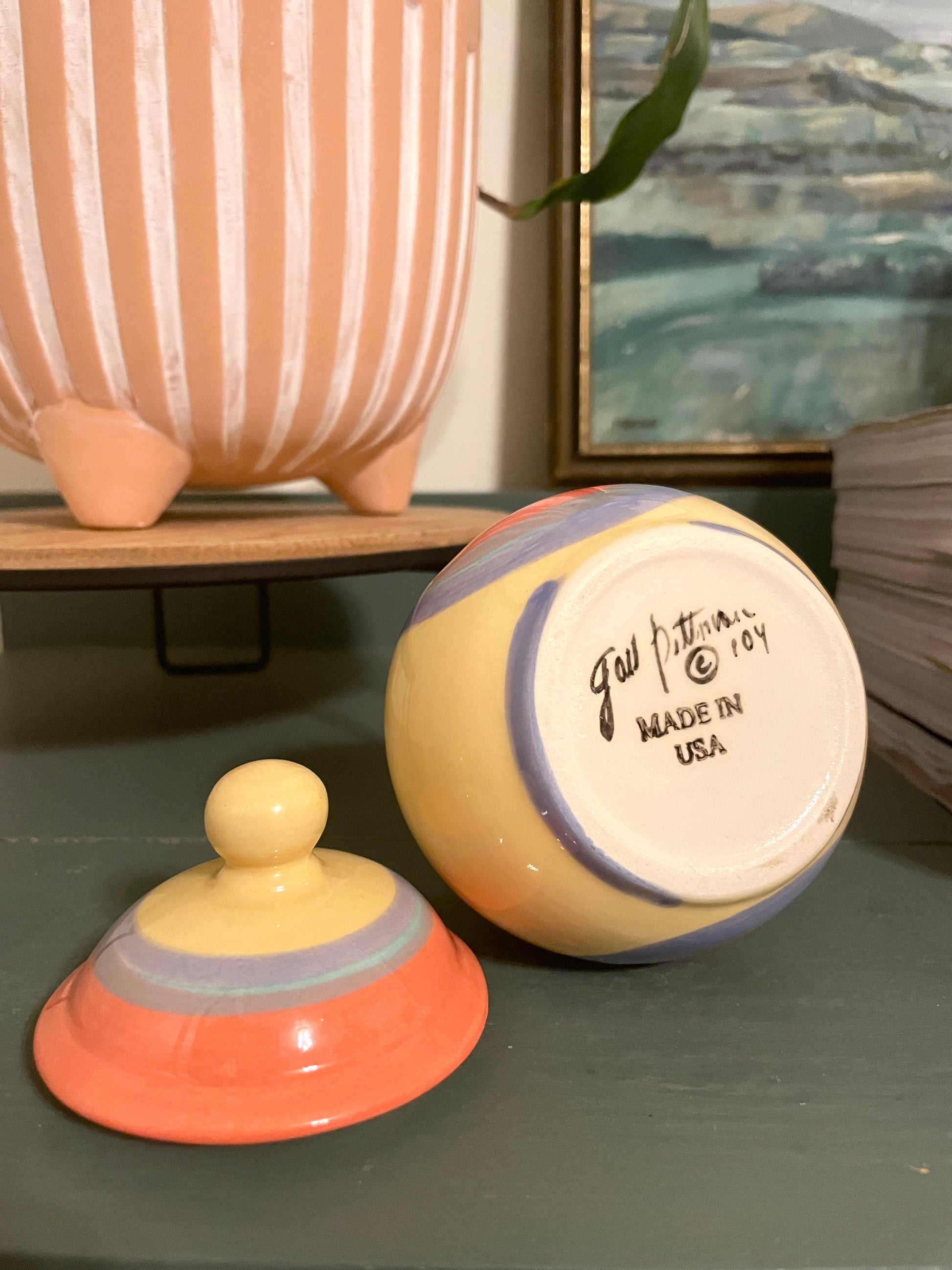 A small hand-painted ceramic lidded container shown on a green tabletop, with the lid off and the base printed with ‘Gail Pittman © 104 MADE IN USA'.