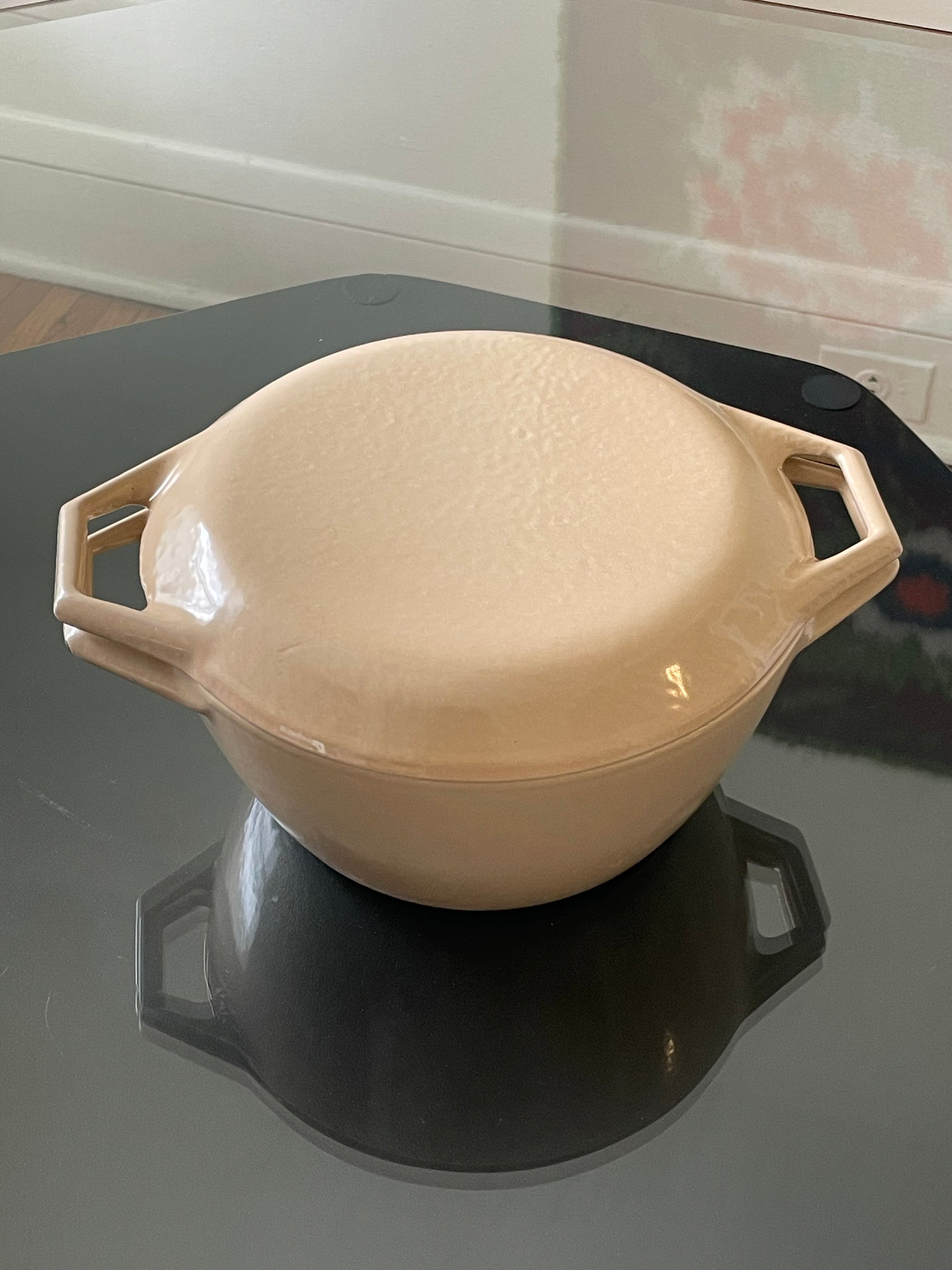 A cream-colored enameled cast iron dutch oven with a matching lid, featuring integrated side handles, resting on a dark reflective surface.