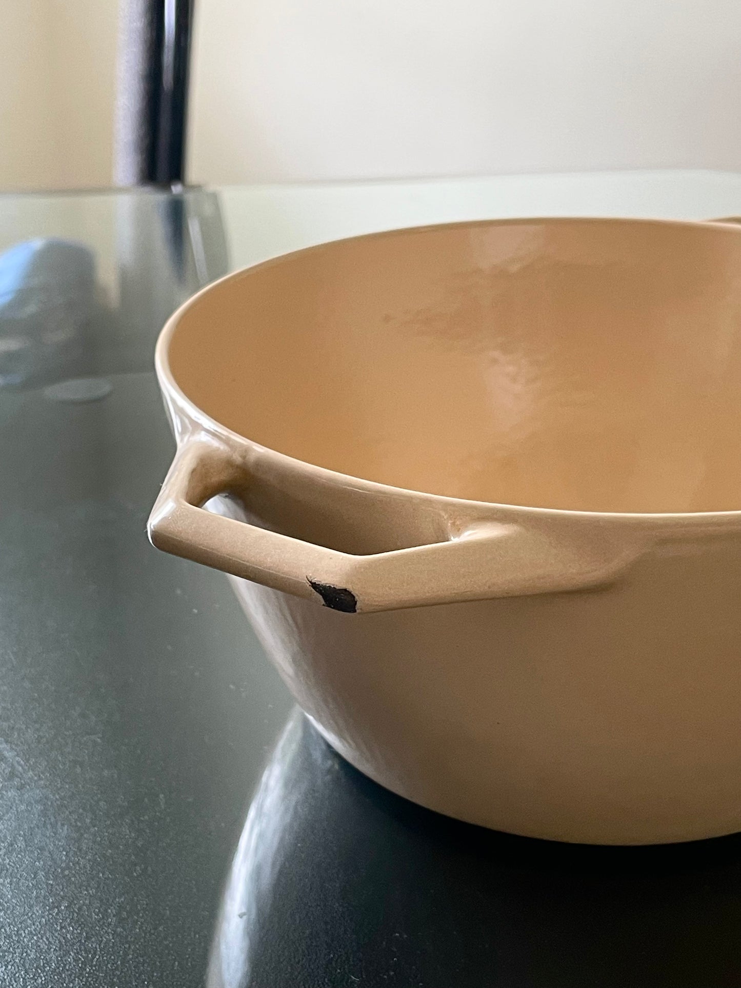 A close-up of a beige enameled cast iron Dutch oven pot with a handle with minor chip to enamel on handle, sitting on a dark reflective countertop.