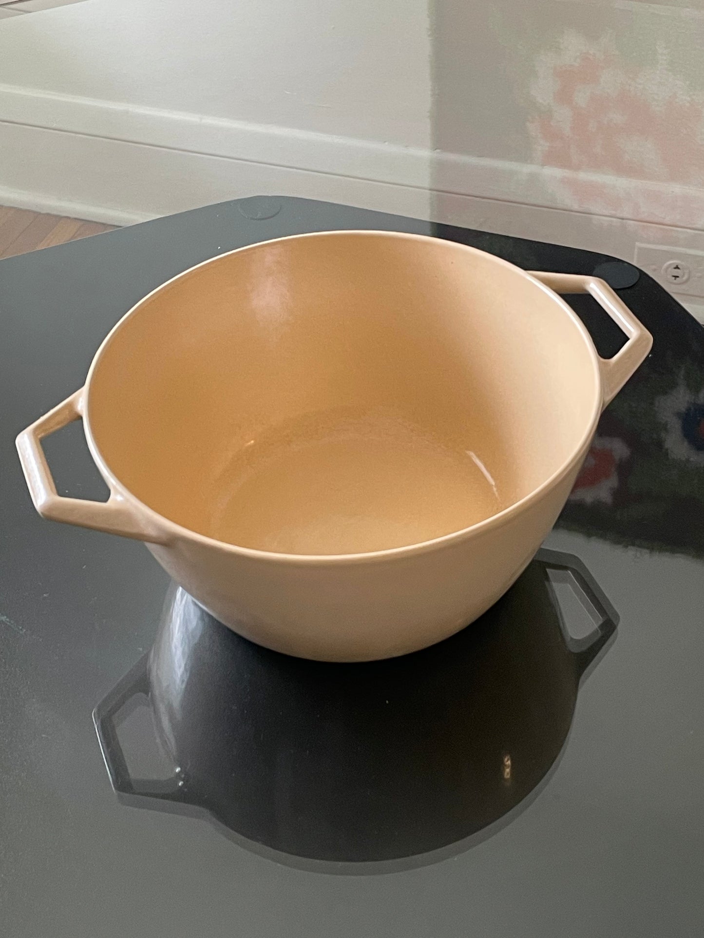 A light beige enameled cast iron Dutch oven with side handles, shown on a black glass table in a home kitchen setting.
