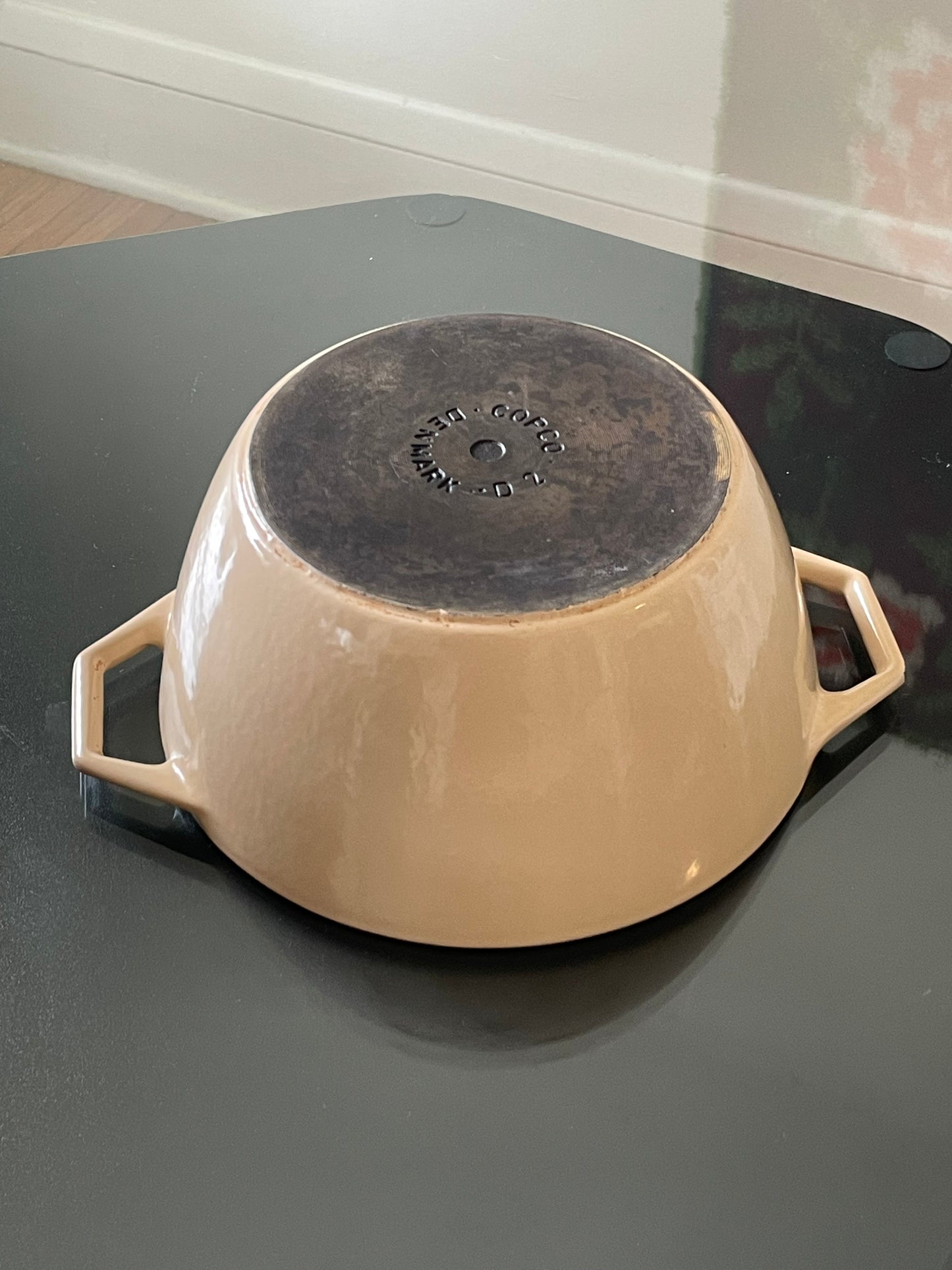 Beige enameled cast iron Dutch oven with side handles, shown upside down on a dark reflective tabletop, with a dark cast-iron bottom base visible. The base reads “Copco Denmark D2”.