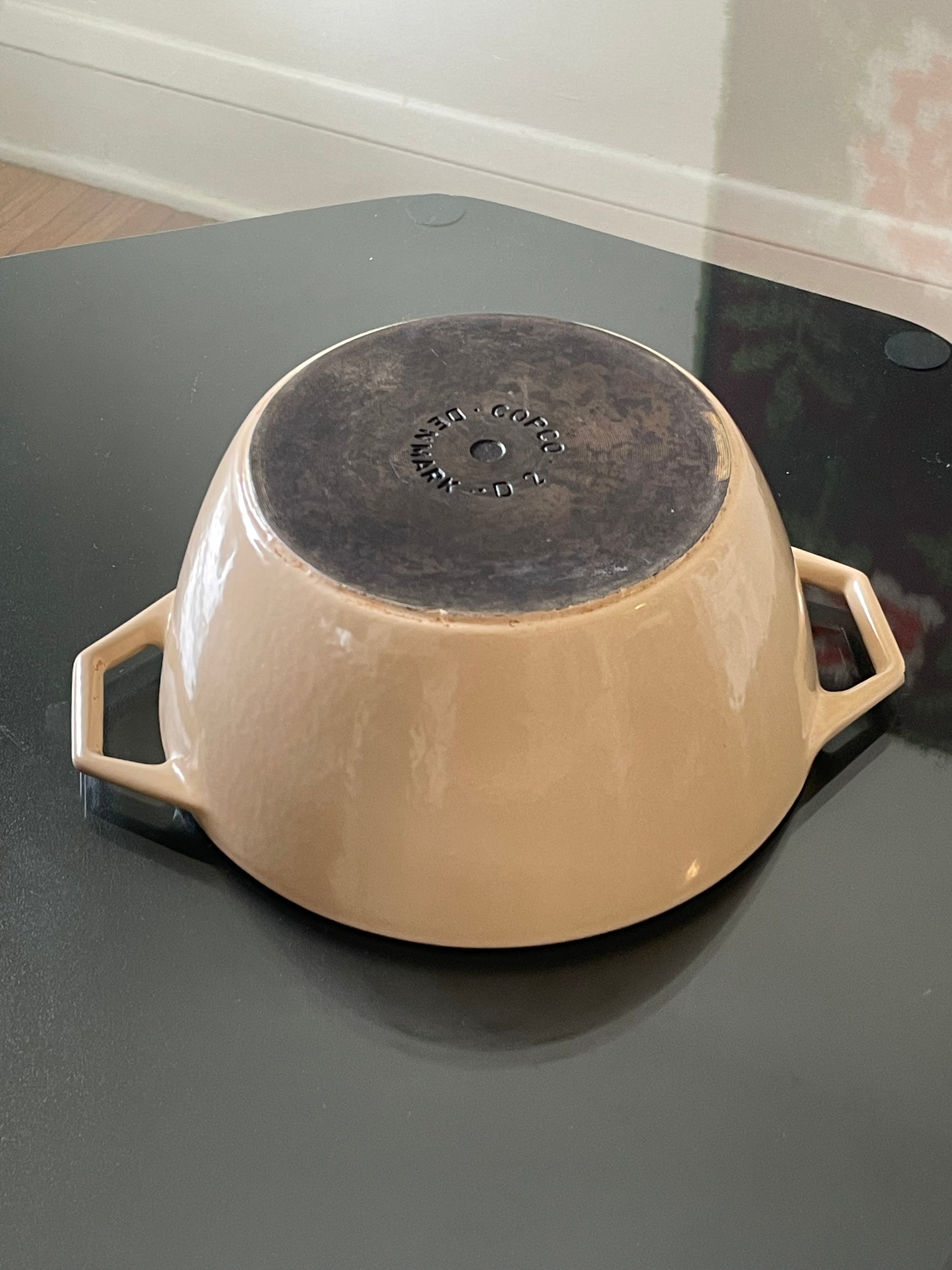 Beige enameled cast iron Dutch oven with side handles, shown upside down on a dark reflective tabletop, with a dark cast-iron bottom base visible. The base reads “Copco Denmark D2”.