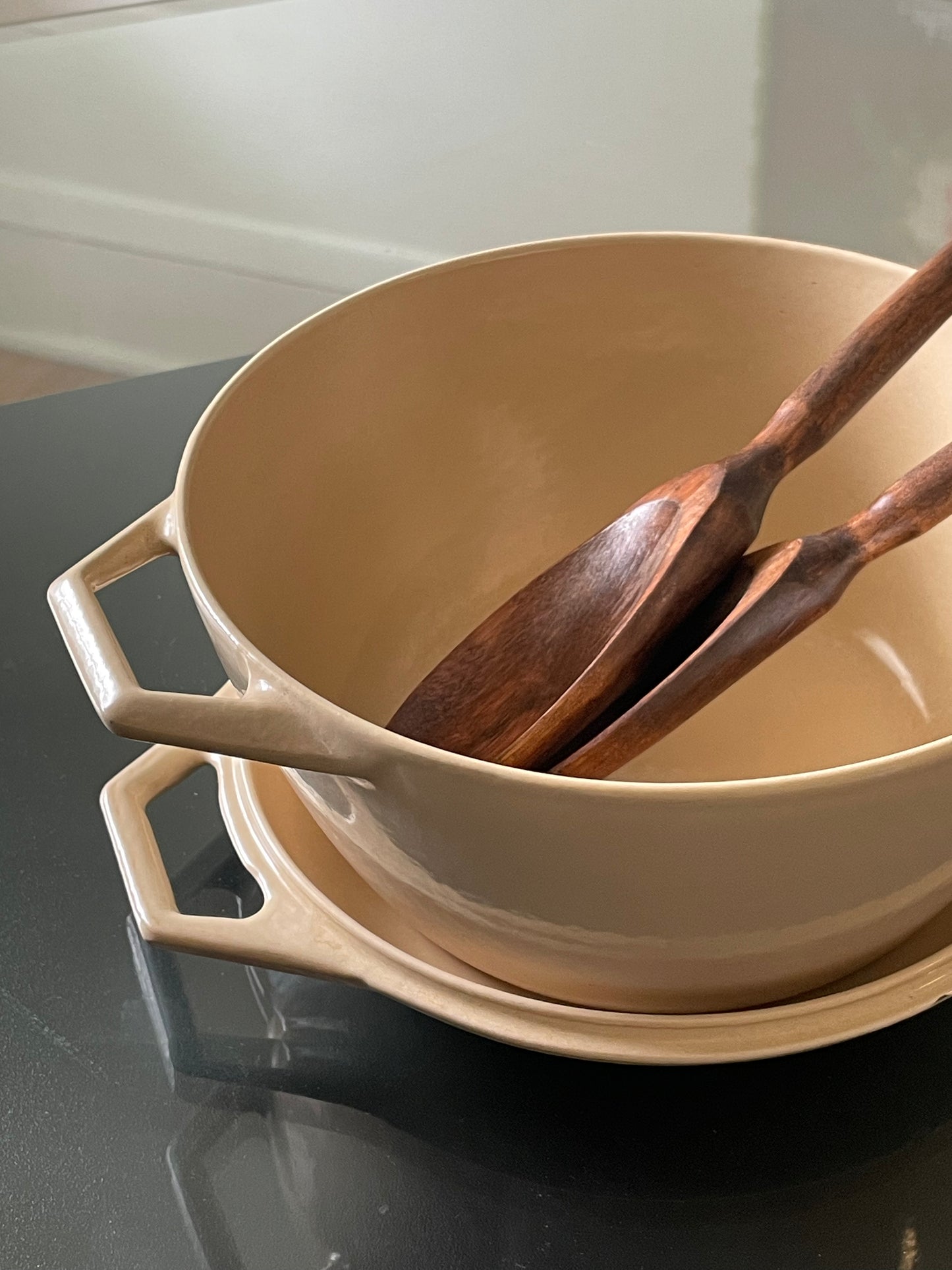 A beige enameled cast iron Dutch oven with side handles, shown stacked on its lid with two wooden serving utensils inside, on a dark reflective surface.