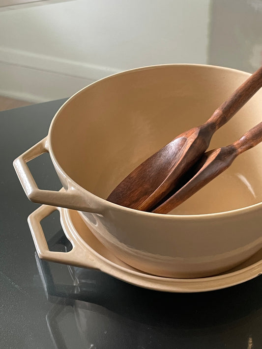 A beige enameled cast iron Dutch oven with side handles, shown stacked on its lid with two wooden serving utensils inside, on a dark reflective surface.