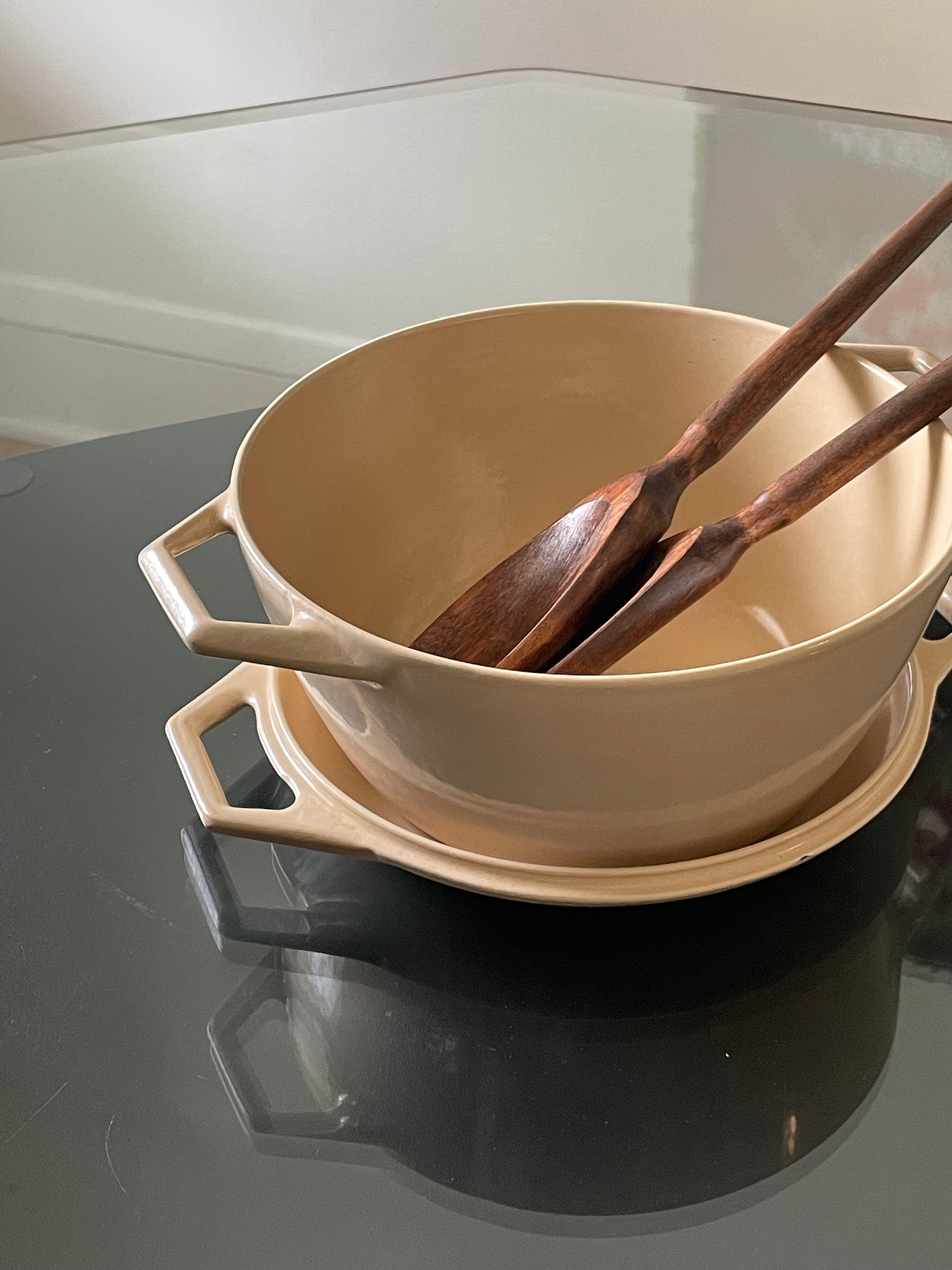 A beige enameled cast iron Dutch oven with side handles, shown stacked on its lid with two wooden serving utensils inside, on a dark reflective surface.