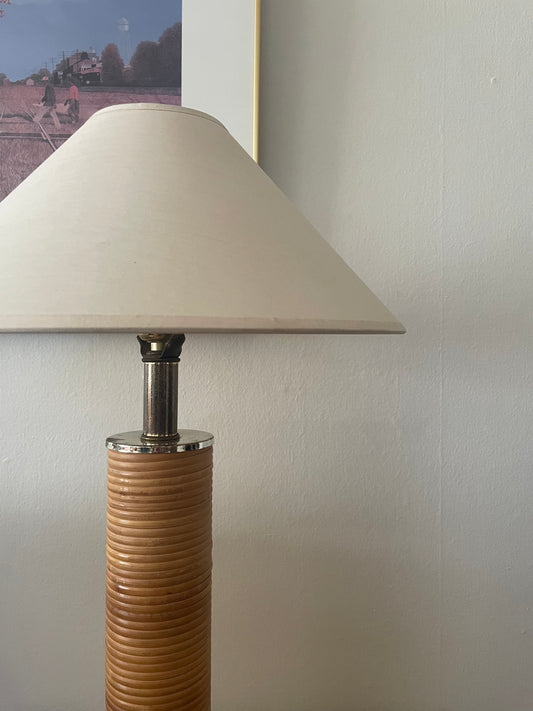 A table lamp with a cream linen taper shade, a silver metal neck, and slender cylindrical base wrapped in light rattan, shown in front of framed artwork against a white wall.