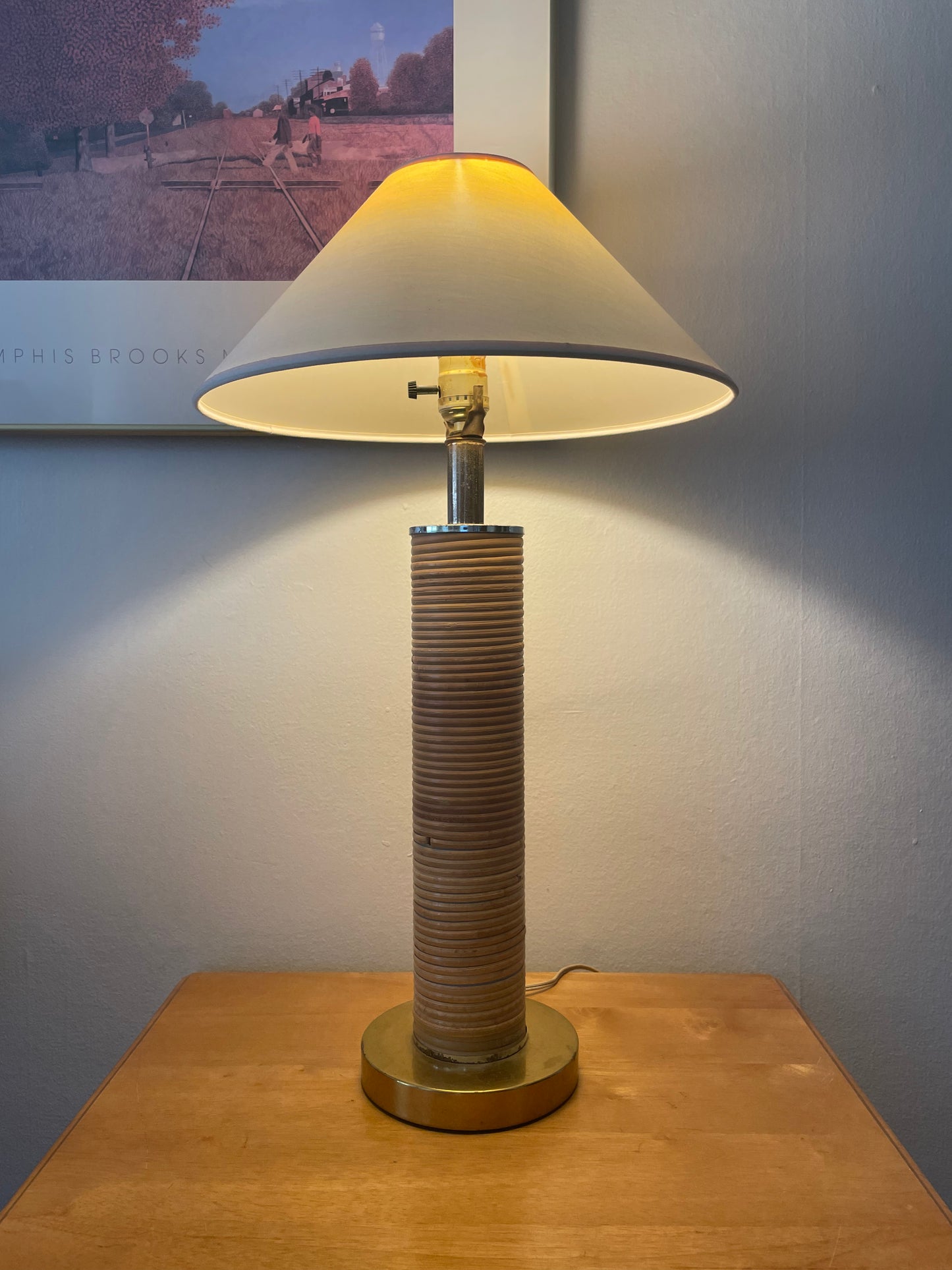 A lit table lamp with a cream linen taper shade, a silver metal neck, and slender cylindrical base wrapped in light rattan on a brass base, sitting on a wood table in front of a white wall with artwork.