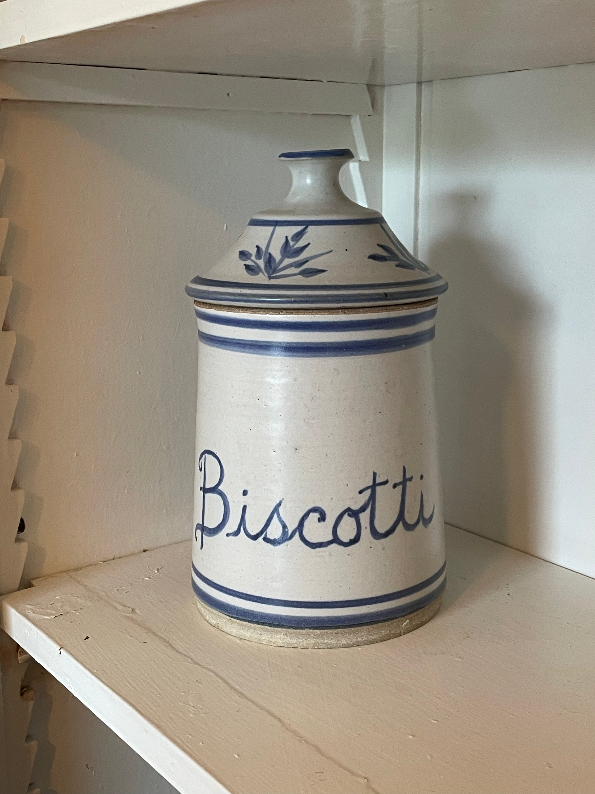A vintage white ceramic biscotti canister with blue stripes and floral decoration, sitting on a painted wooden shelf.